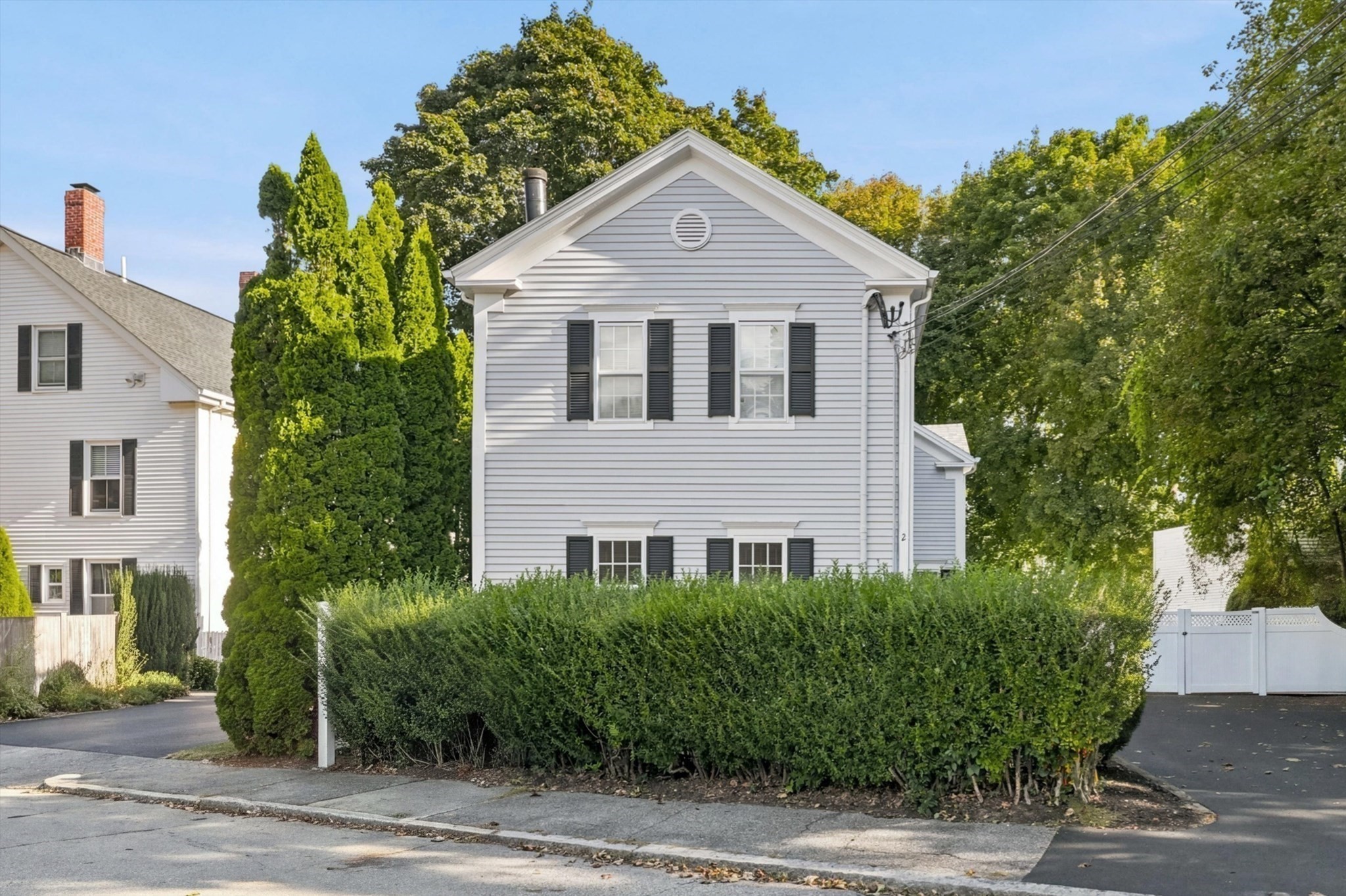 Light gray two-story home with black shutters and a tall hedge along the sidewalk.