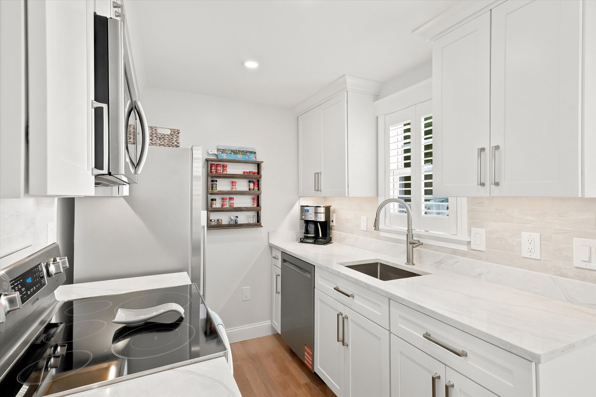 Bright white kitchen with stone counters, stainless steel appliances, and a sink beneath shuttered windows.
