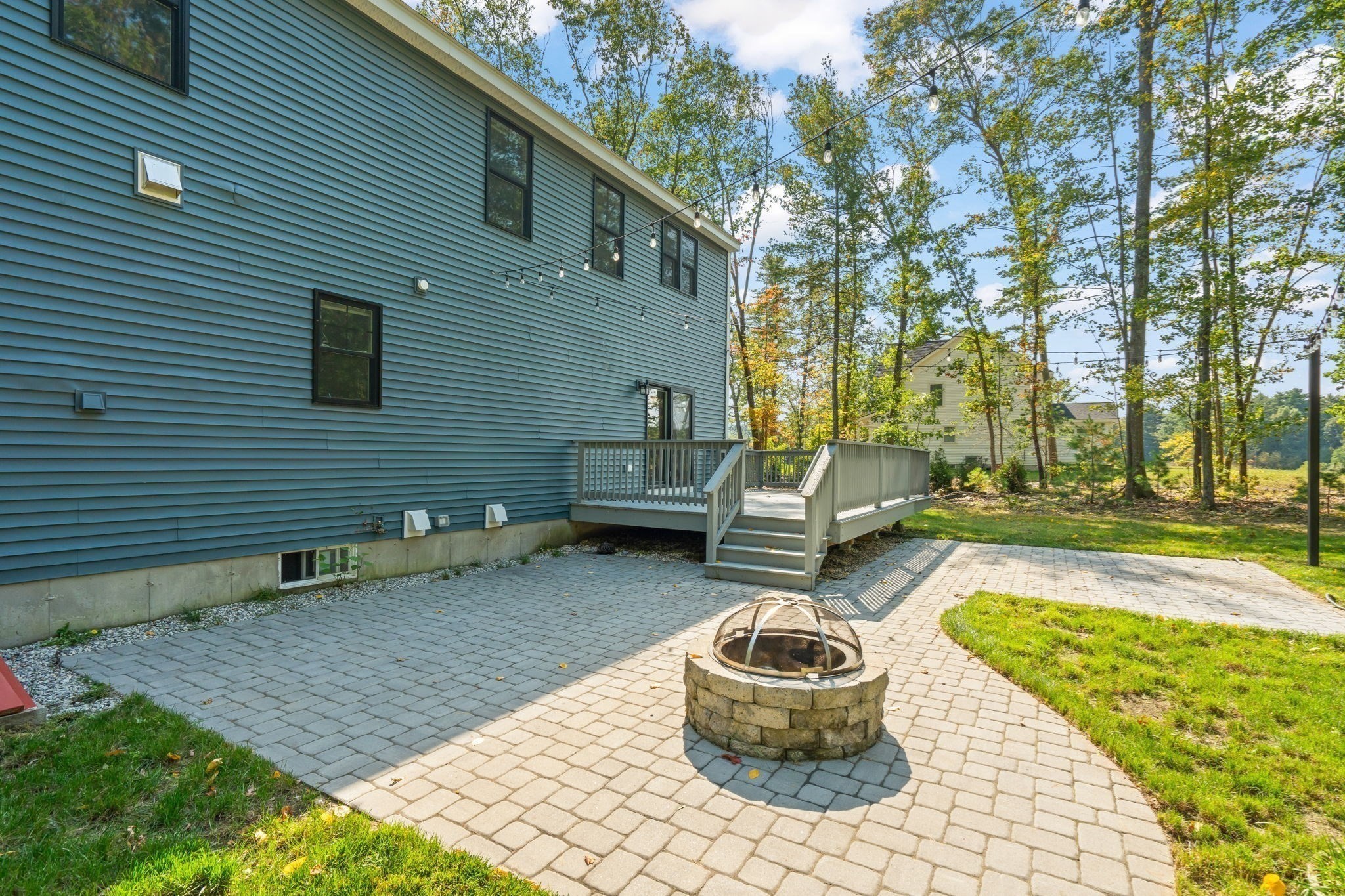 Home’s paved backyard patio with a stone fire pit, raised deck, and overhead string lights.