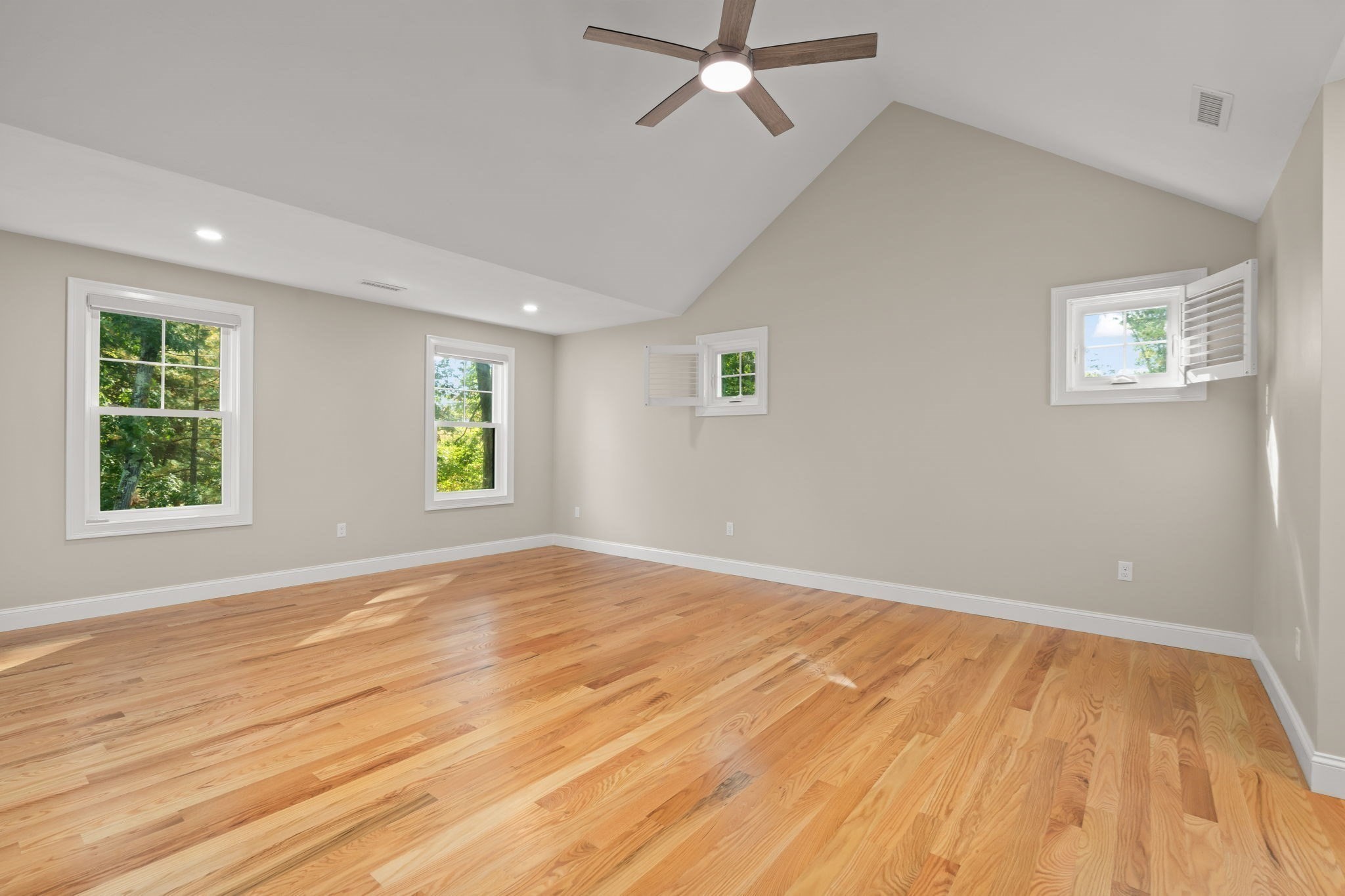 Empty room with polished hardwood floors, vaulted ceiling, ceiling fan, and windows with plantation shutters.