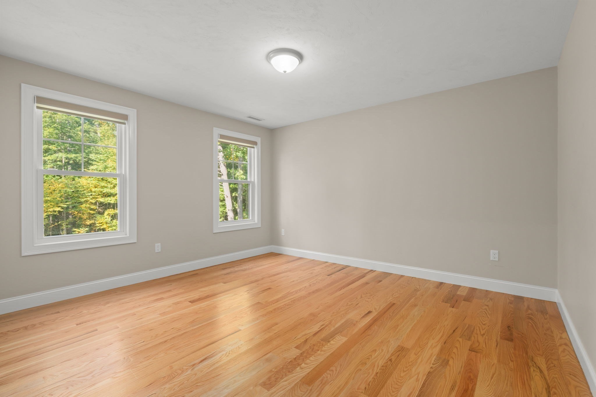 Empty bedroom with two tree-facing windows and polished hardwood floors.