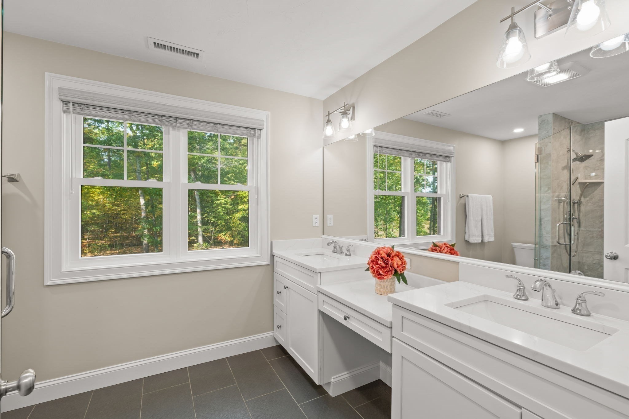 Bathroom with double white vanities, a large mirror, and windows overlooking leafy trees.