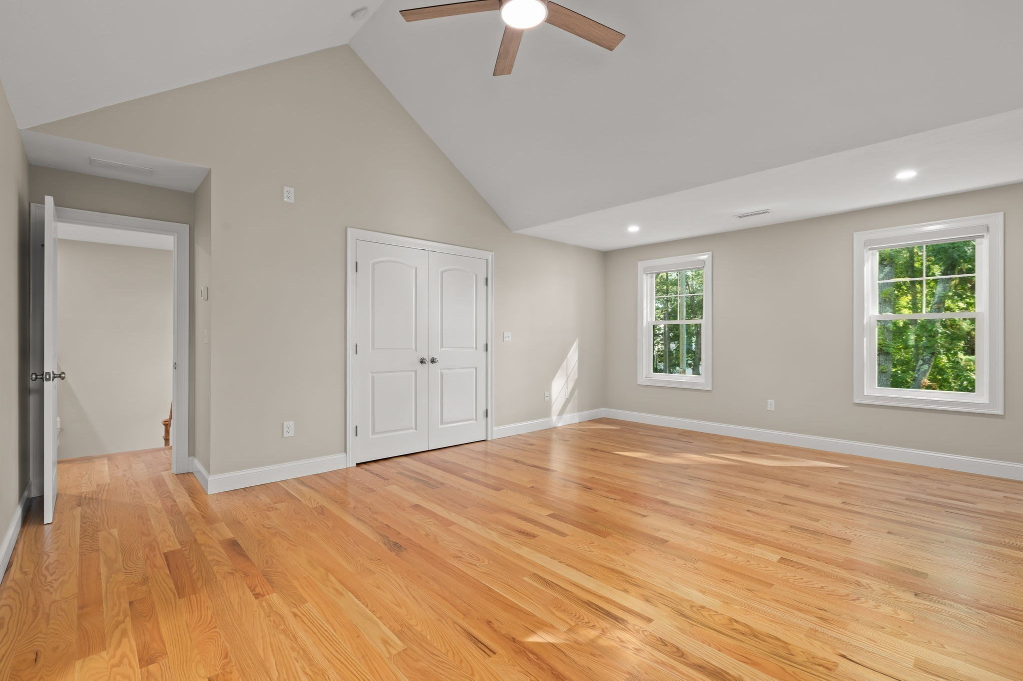 Empty bedroom with hardwood floors, vaulted ceiling, double closet doors, and windows overlooking trees.