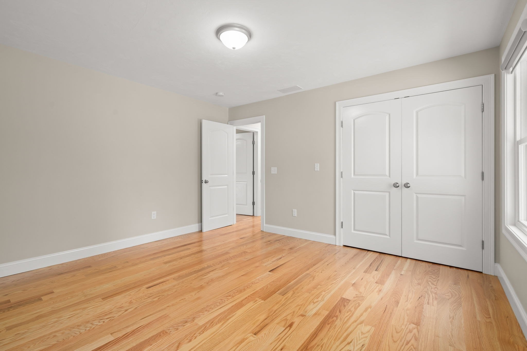 Empty bedroom with hardwood floors, white double closet doors, and a window.