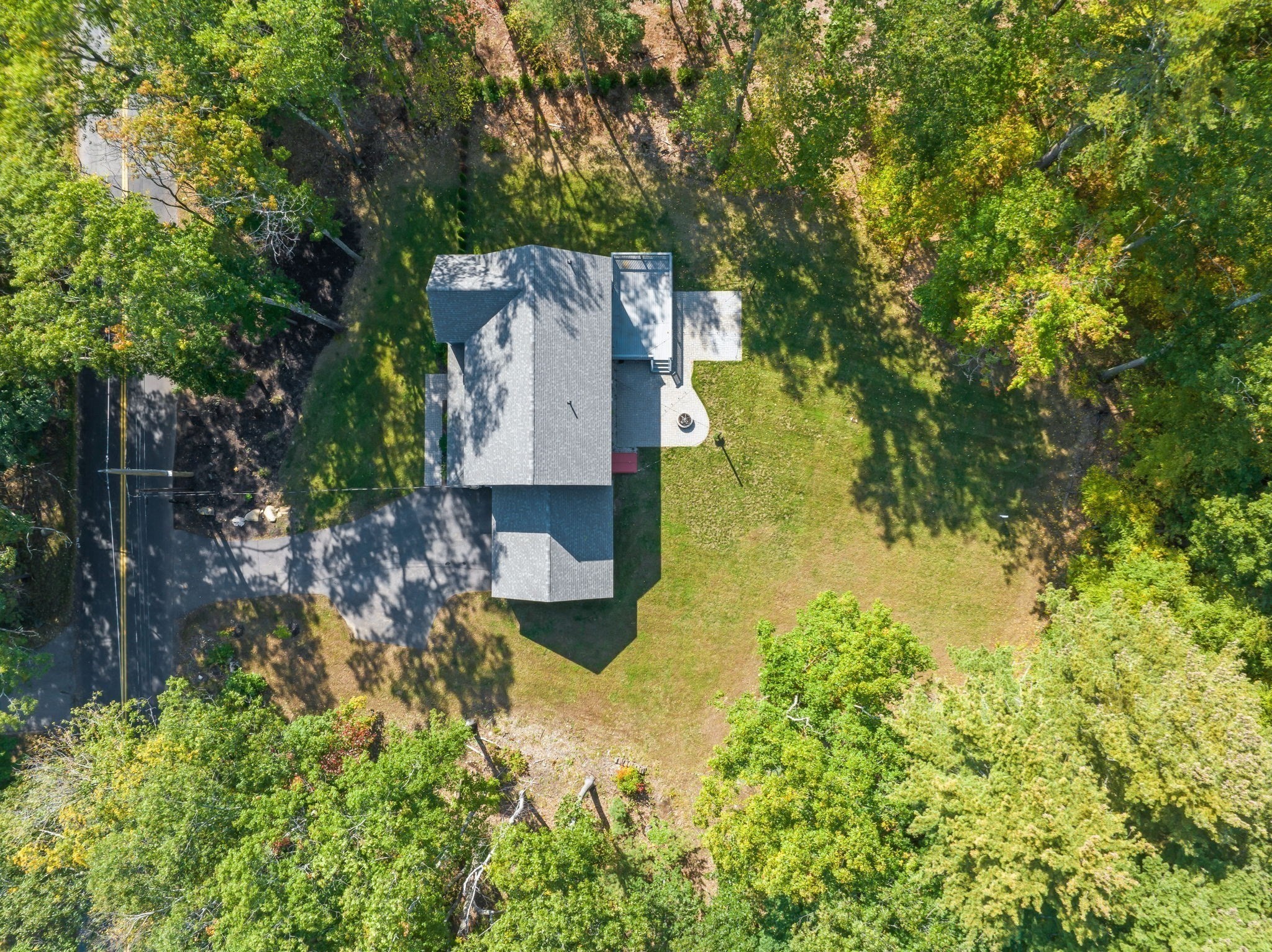 Aerial view of a house on a wooded lot, with a broad lawn, paved driveway, and small rear patio.