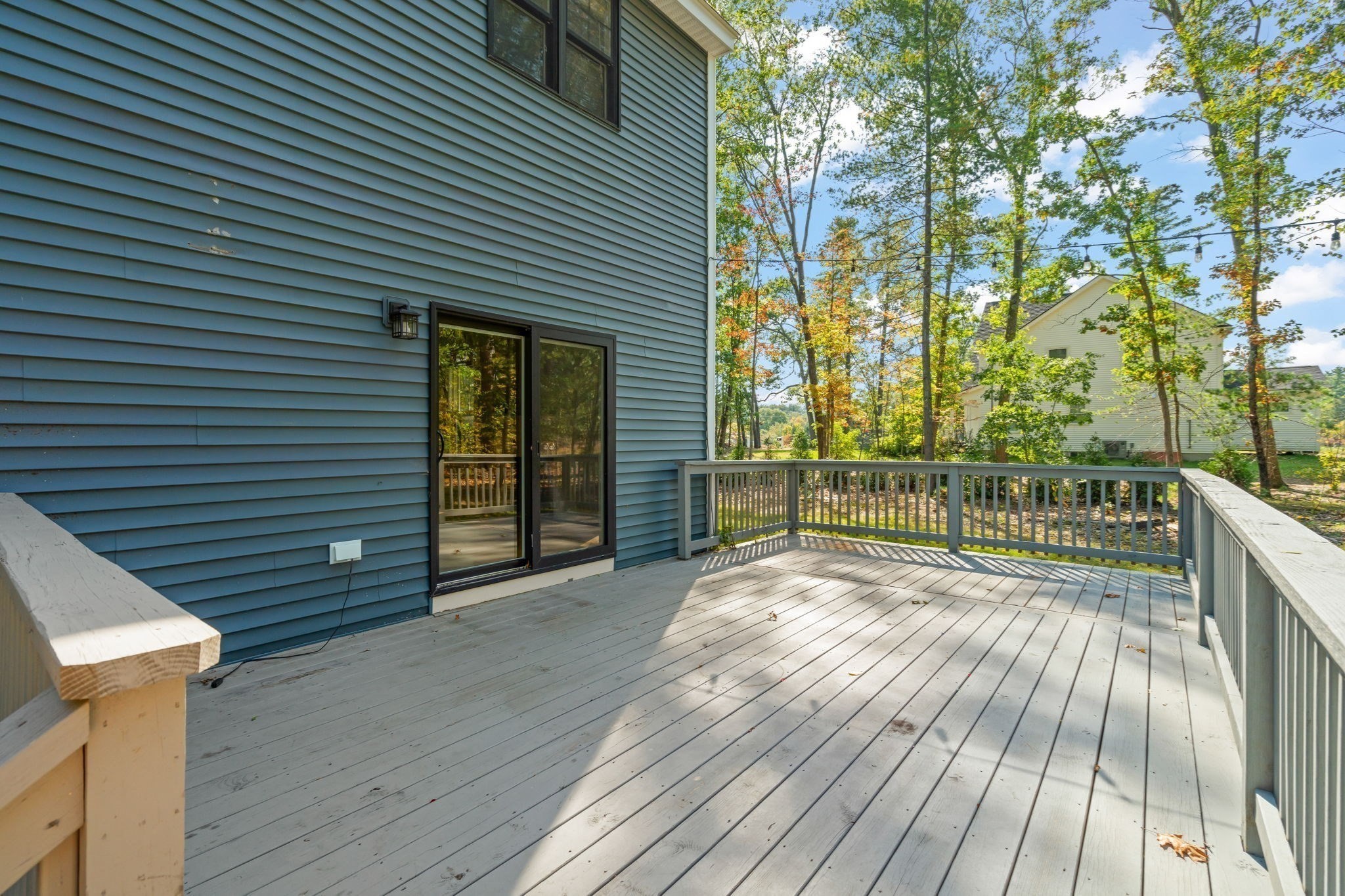 Spacious gray deck beside a blue-sided house, with railings and trees beyond.