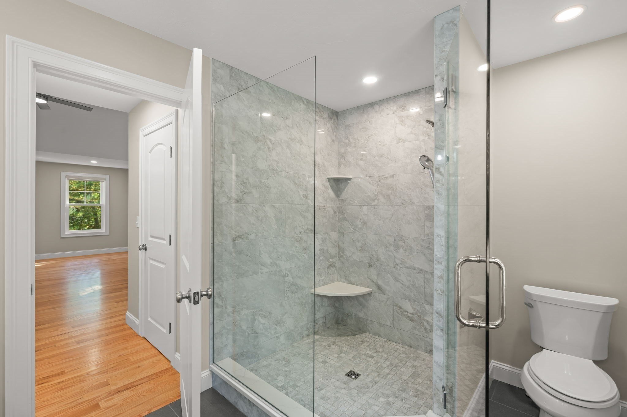 Bathroom with a glass-enclosed gray-tiled walk-in shower and white toilet beside an open doorway.