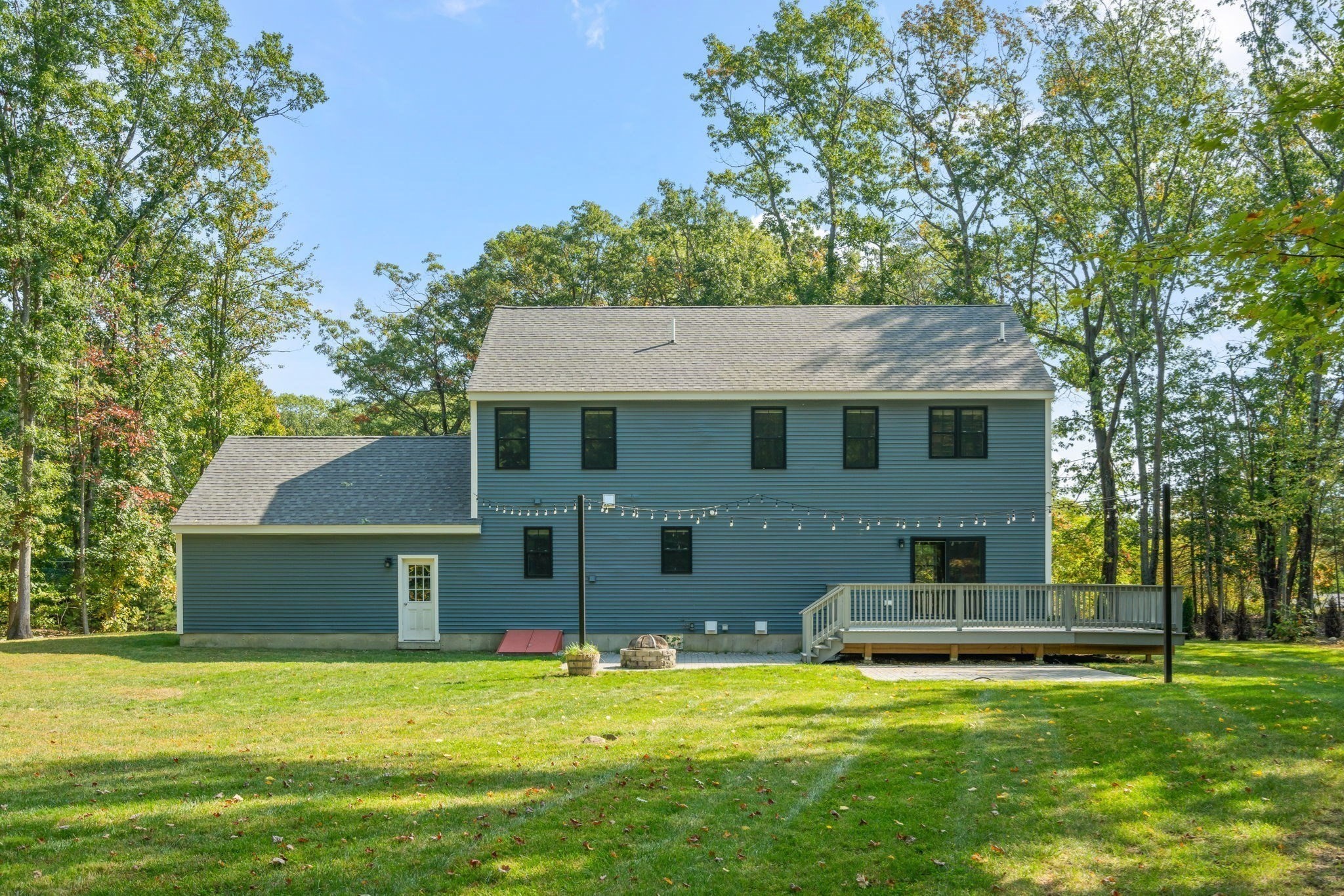 Blue two-story house with a raised deck, string lights, and fire pit in a wooded yard.