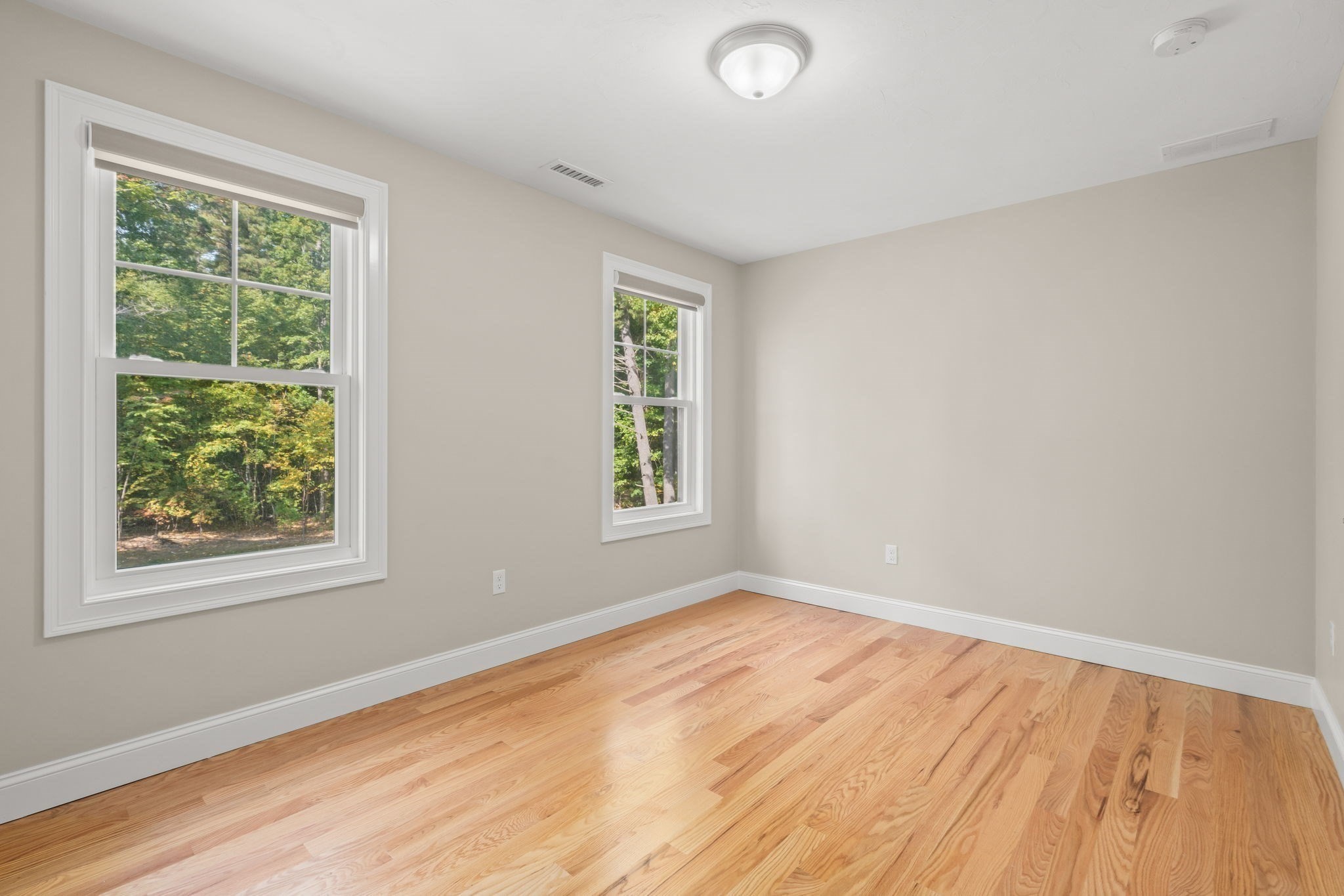 Empty room with hardwood floors, beige walls, and two windows overlooking trees.
