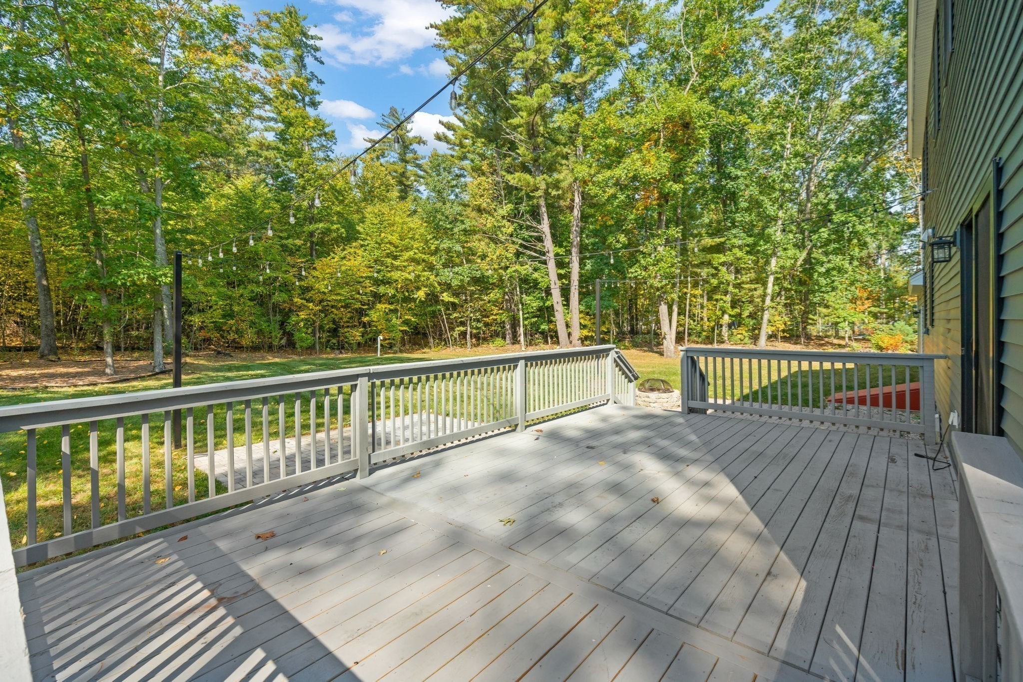 Spacious gray deck overlooking a wooded backyard, with railings and overhead string lights.