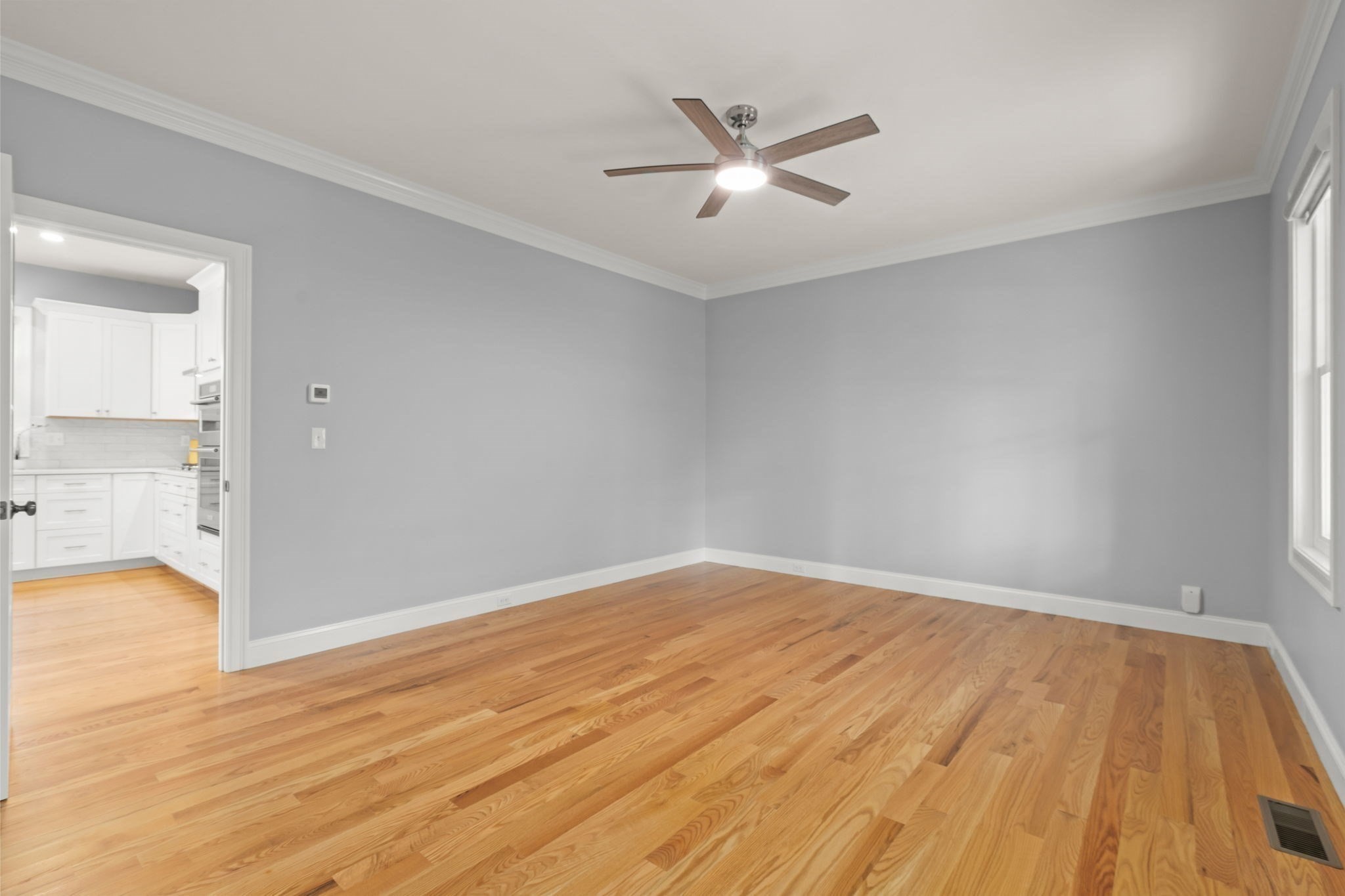 Empty room with hardwood floors, gray walls, a ceiling fan, and an adjoining kitchen.