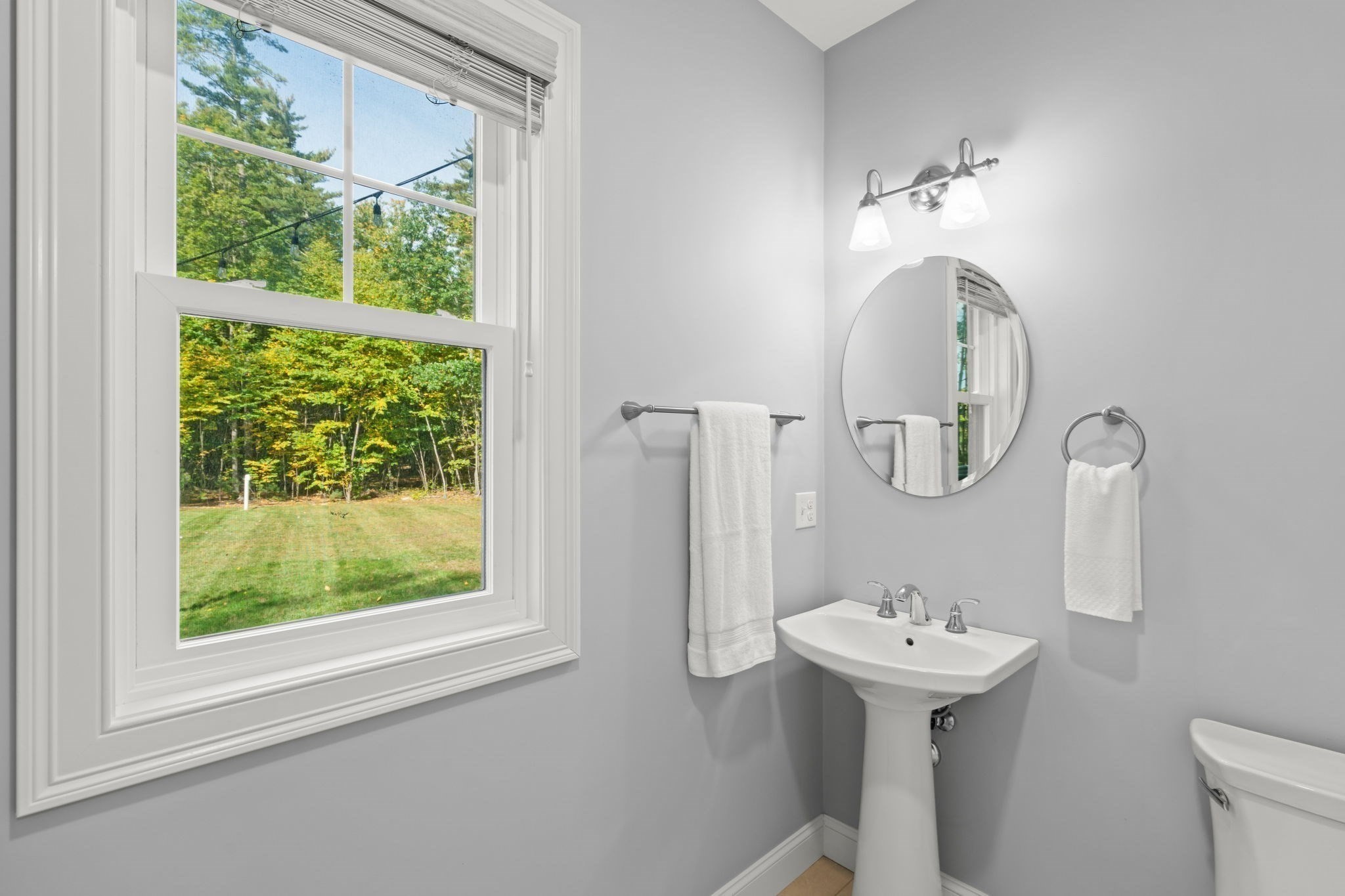 Bathroom with a pedestal sink and round mirror beside a large window overlooking a wooded yard.