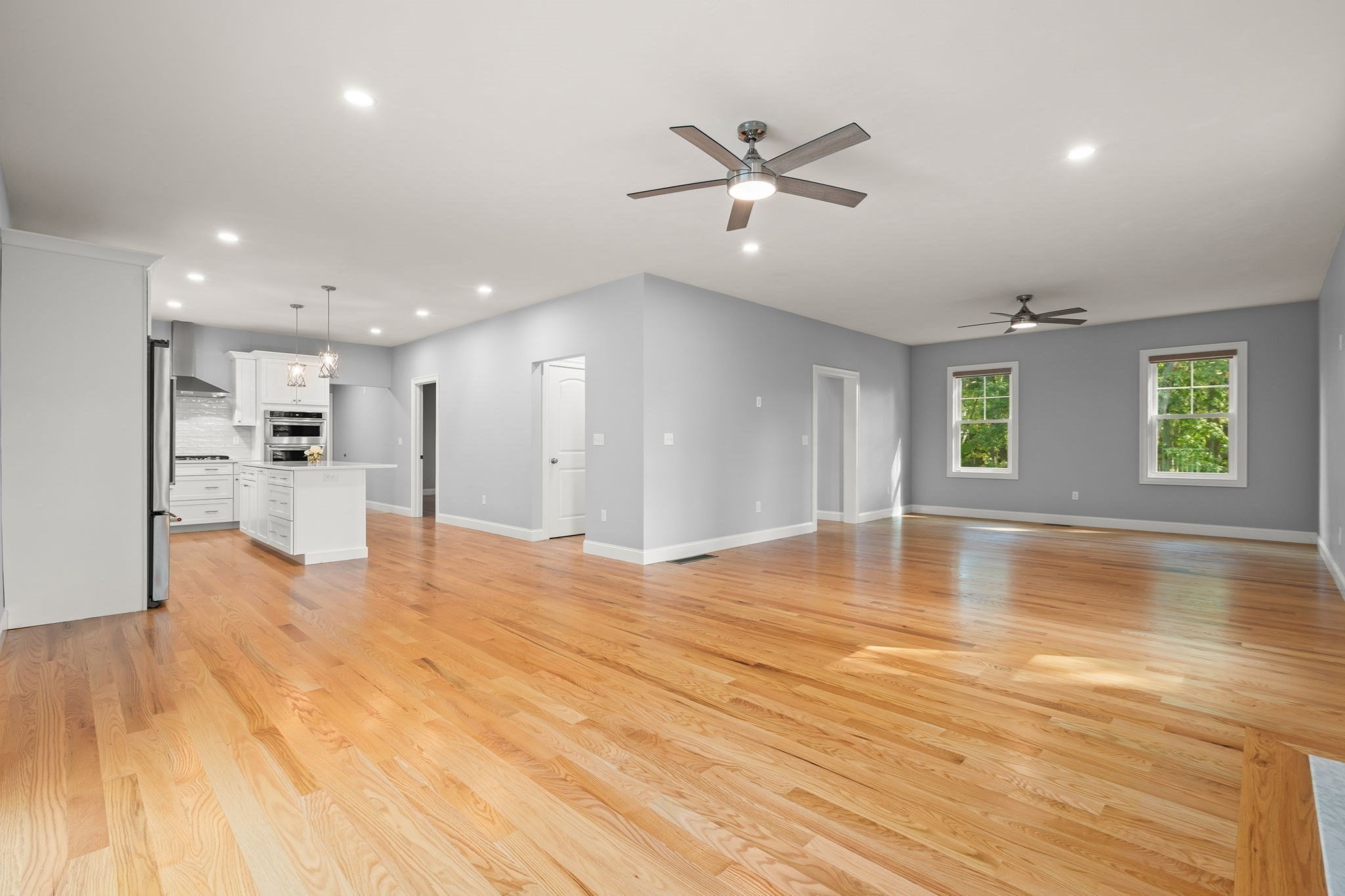 Open-plan living area and white kitchen with hardwood floors, gray walls, ceiling fans, and two windows.
