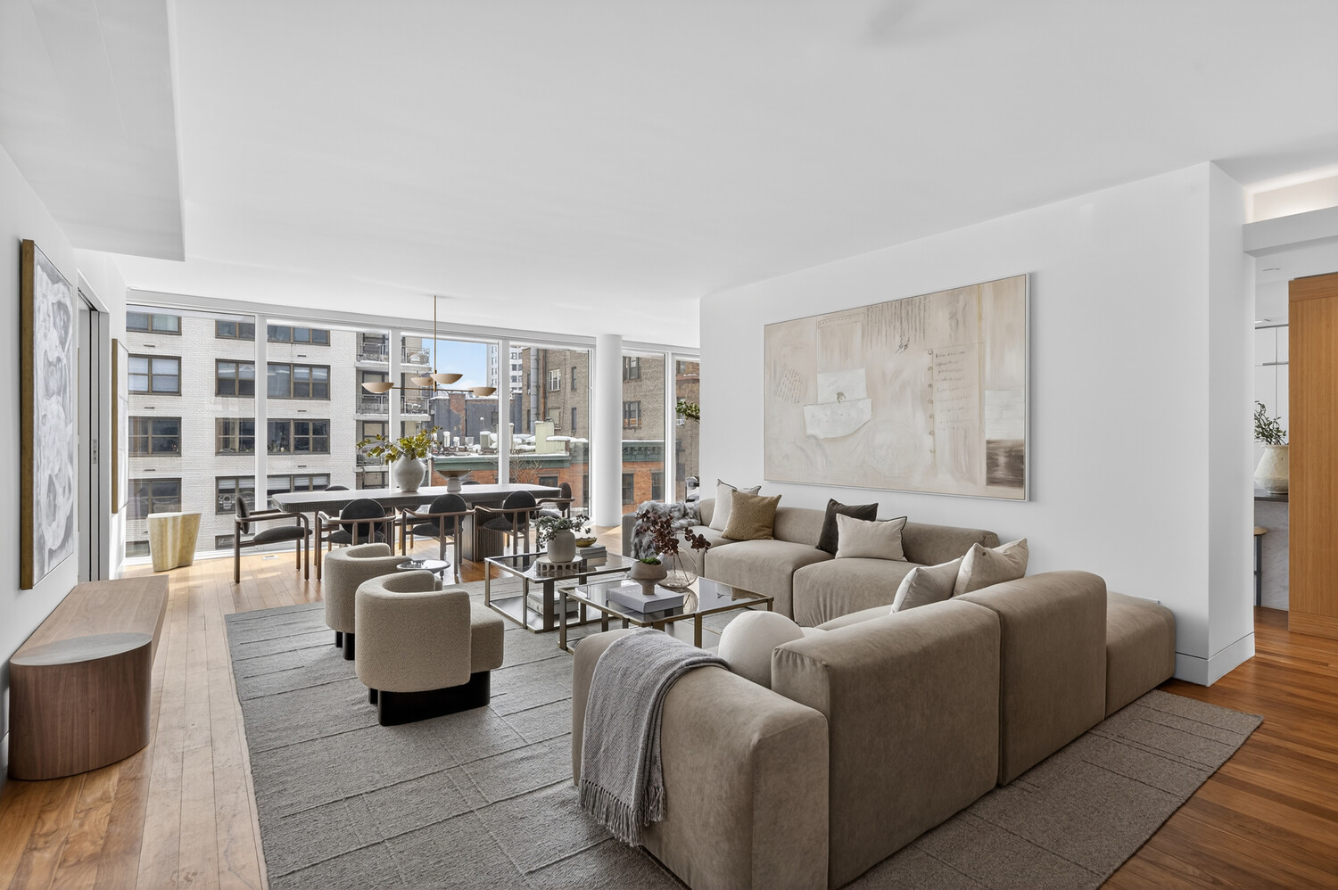 Open-plan living and dining room with a large sectional sofa and floor-to-ceiling windows overlooking city buildings.
