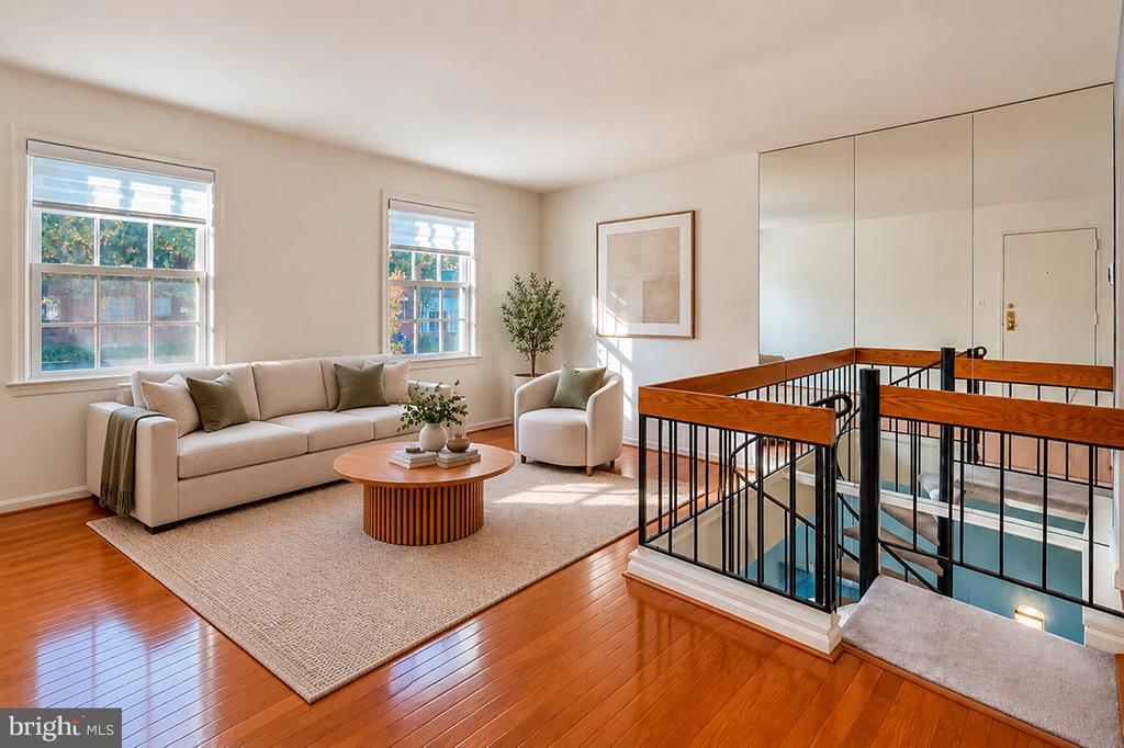 Staged living area with beige seating, polished wood floors, and a railing around an open stairwell.