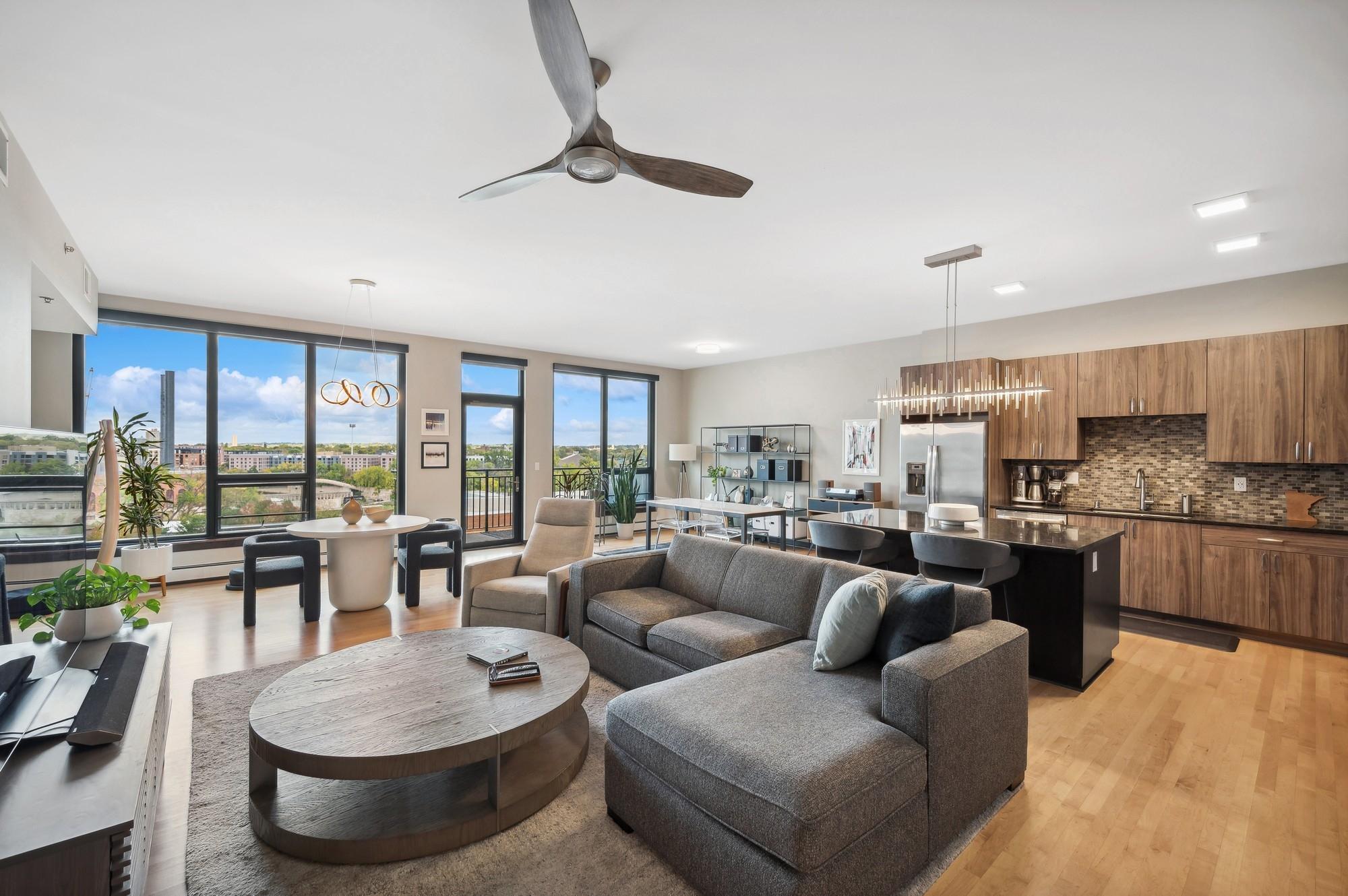 Open-plan living room and kitchen with a gray sectional, wood cabinetry, and large windows overlooking city buildings.
