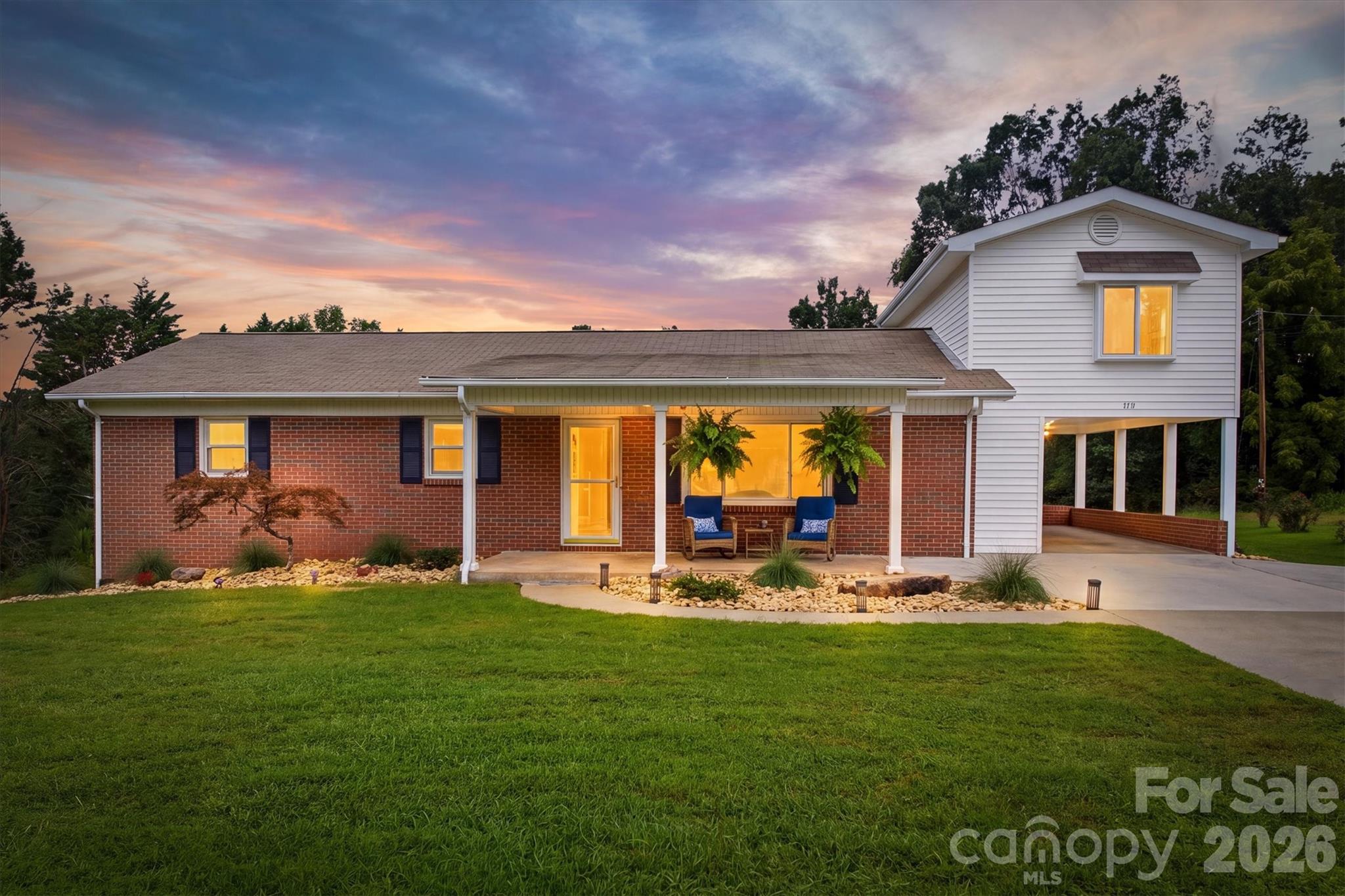 Brick-and-white house with a covered porch, blue chairs, and attached carport at dusk.