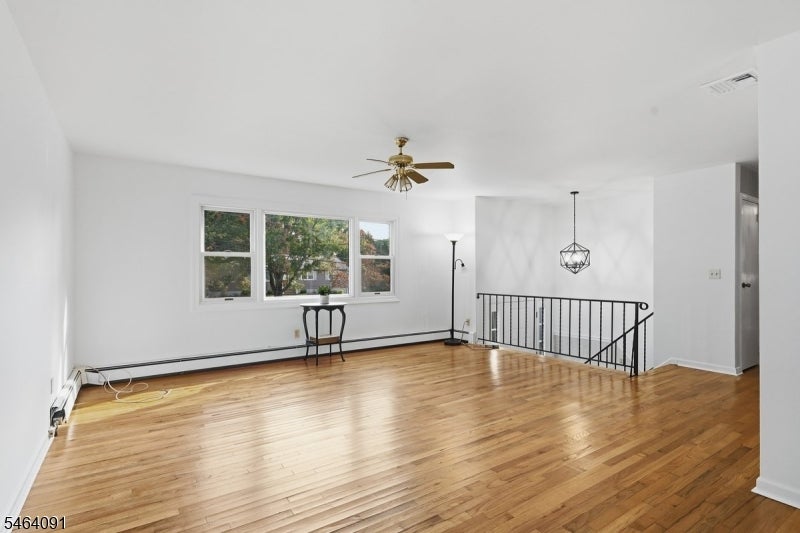 Mostly empty living room with polished hardwood floors, large windows, ceiling fan, and open stairwell railing.
