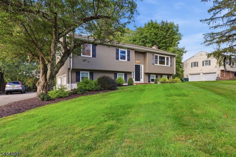 Taupe split-level home with dark shutters, a broad sloping lawn, and a large shade tree by the driveway.