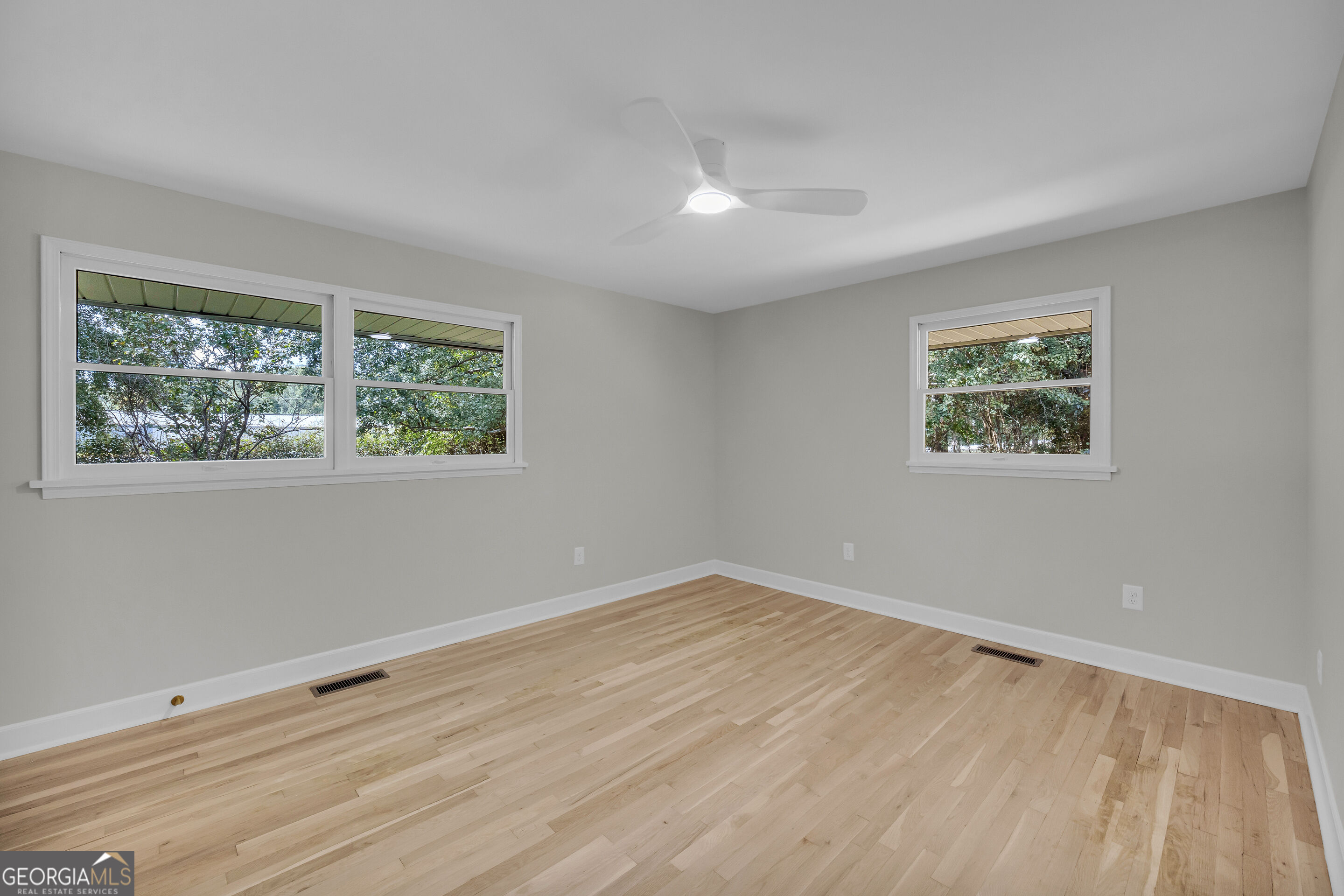 Empty bedroom with light hardwood floors, three windows, and a white ceiling fan.
