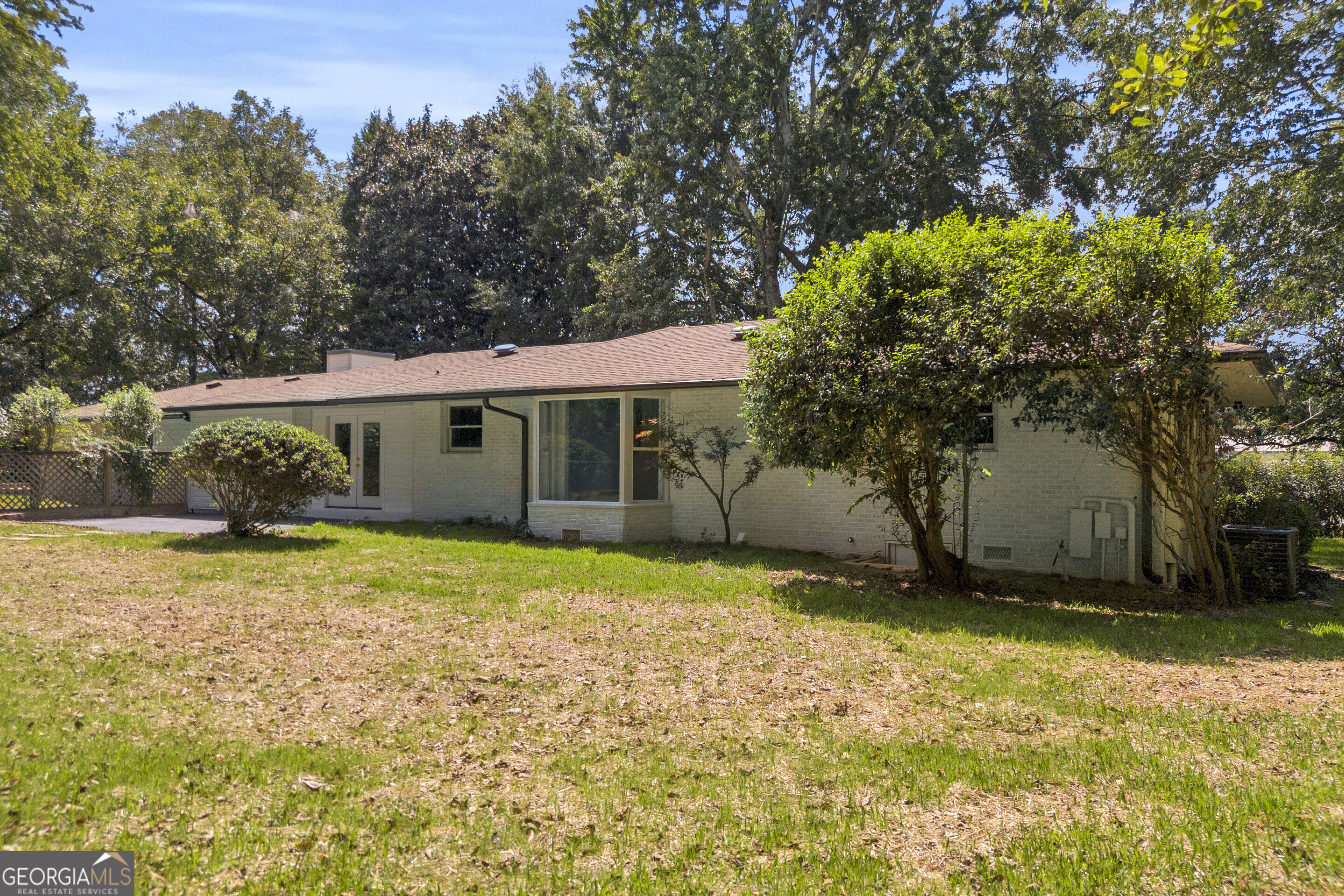 Single-story brick house with a low roof, shaded by mature trees beside a patchy lawn.