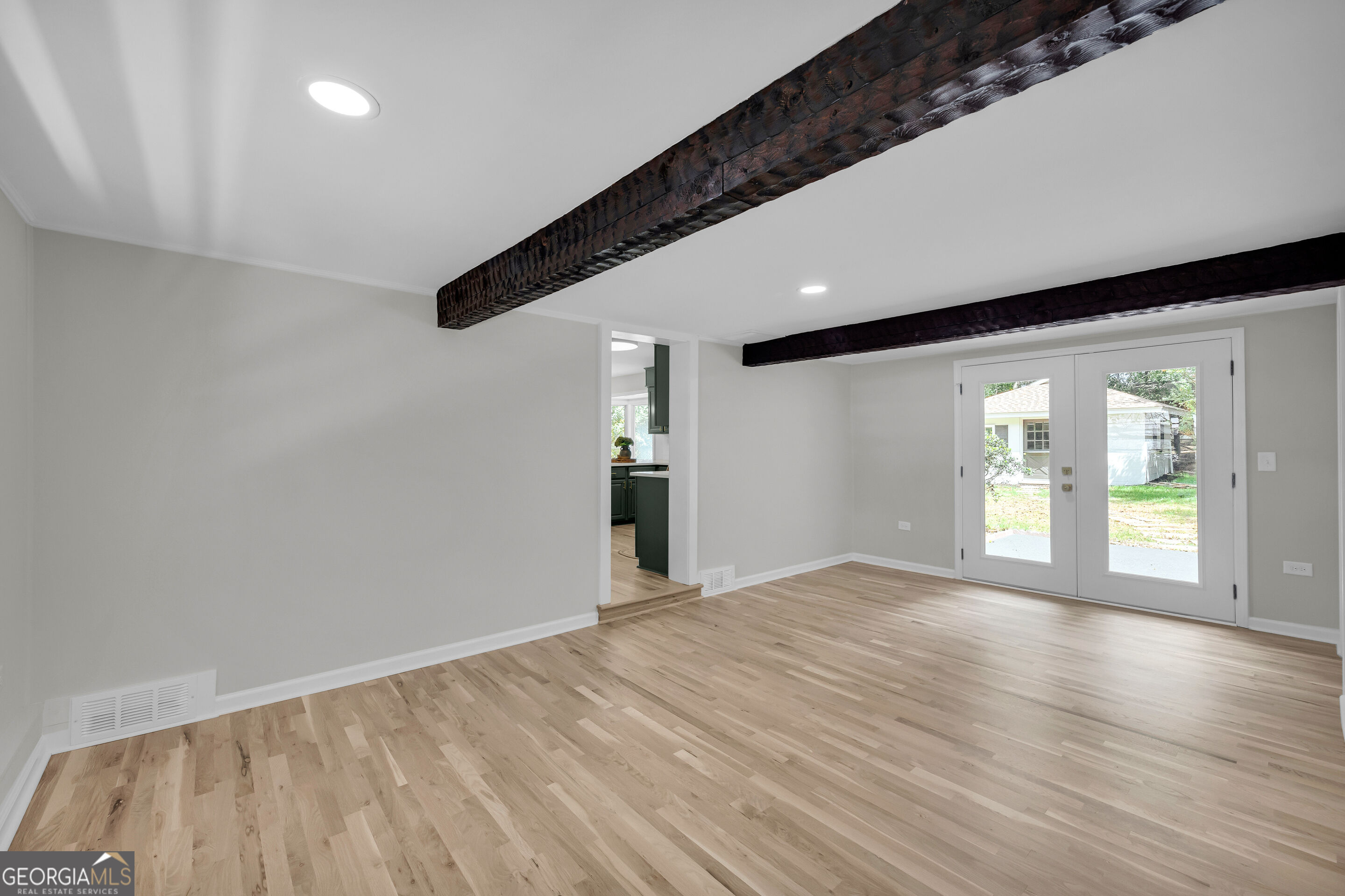 Empty room with light hardwood floors, dark exposed ceiling beams, and glass double doors to the yard.