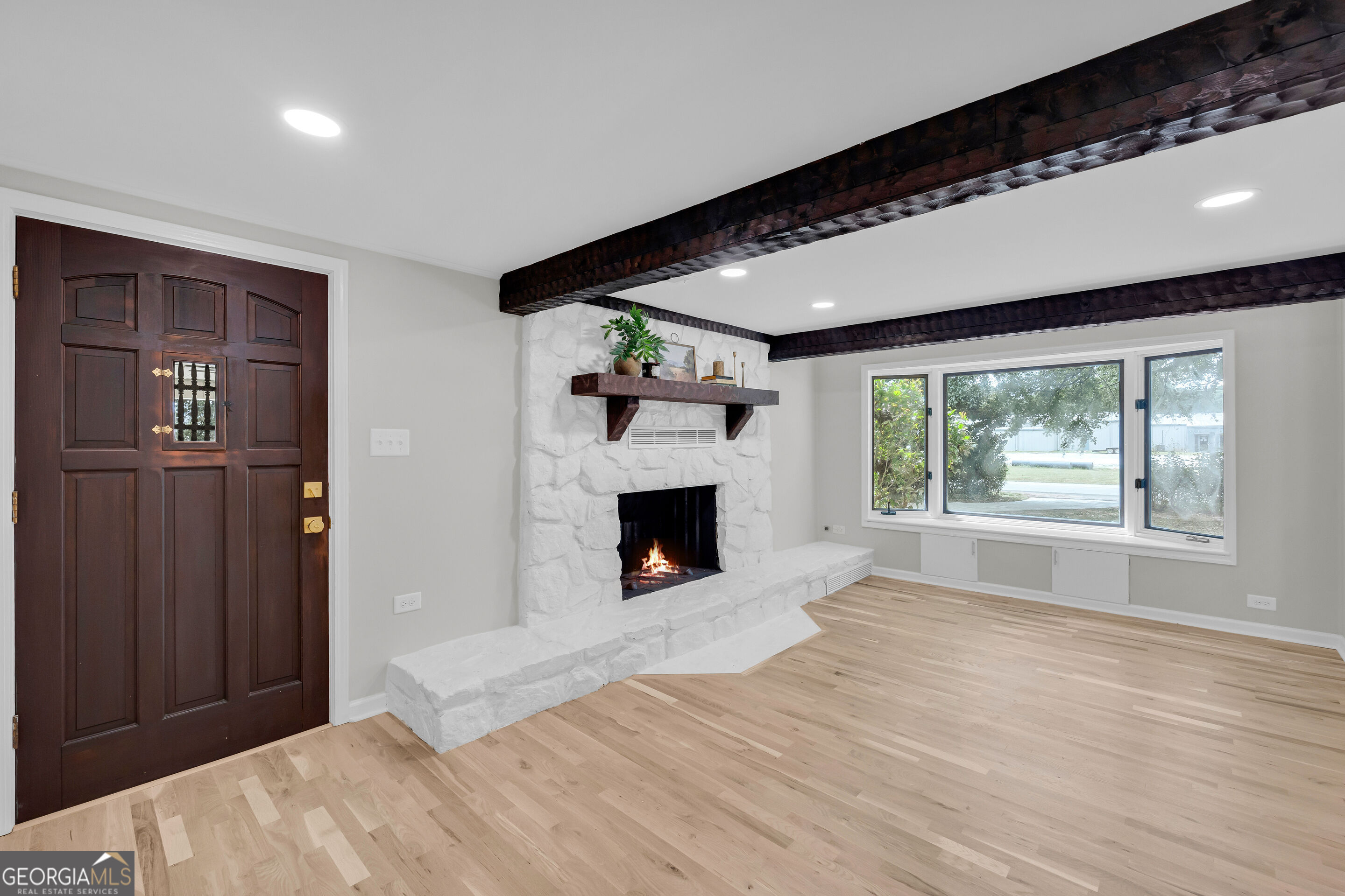 Empty living room with a white stone fireplace, dark ceiling beams, and light hardwood floors.