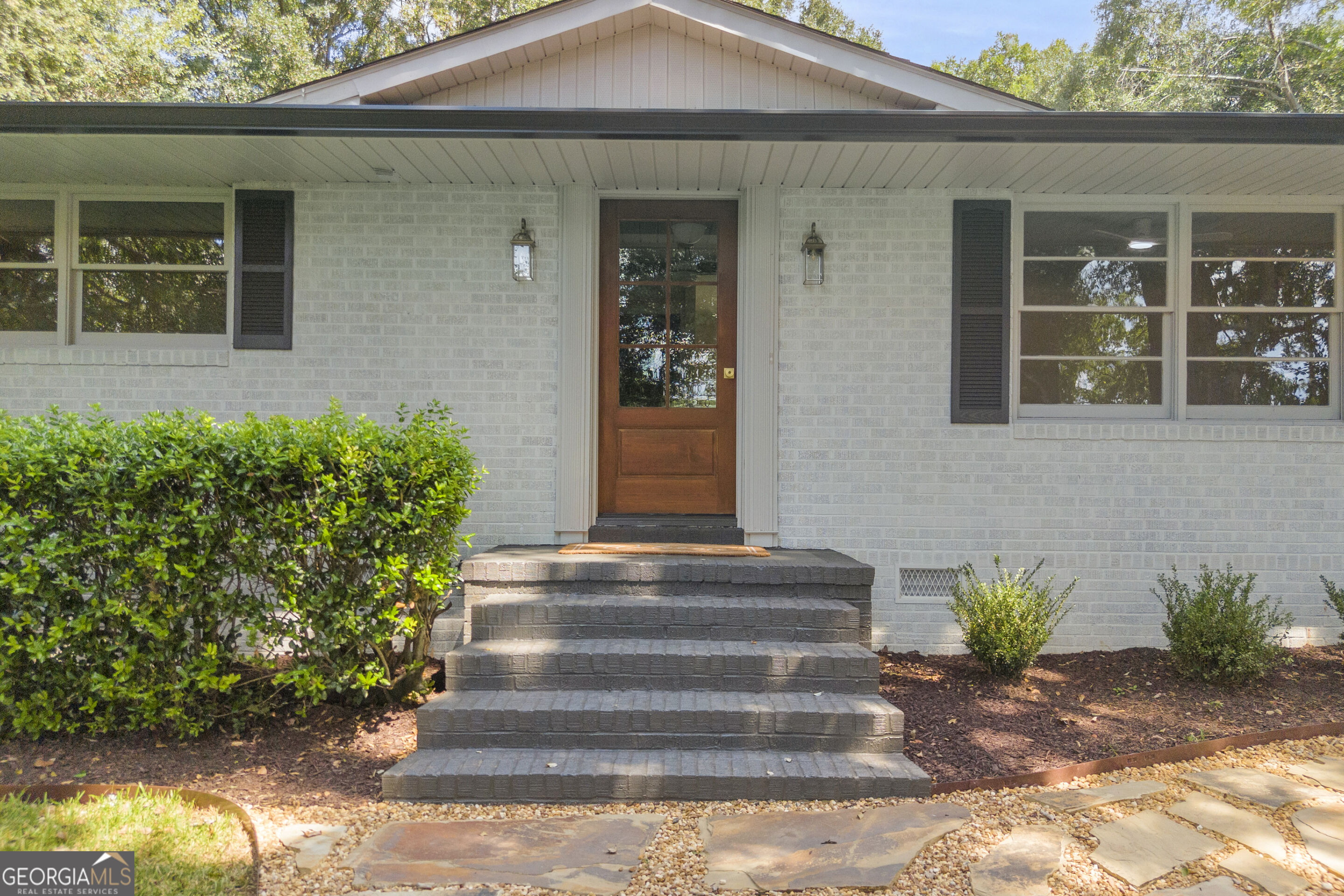 White brick home entrance with a wood-and-glass door, broad brick steps, shrubs, and a stone path.