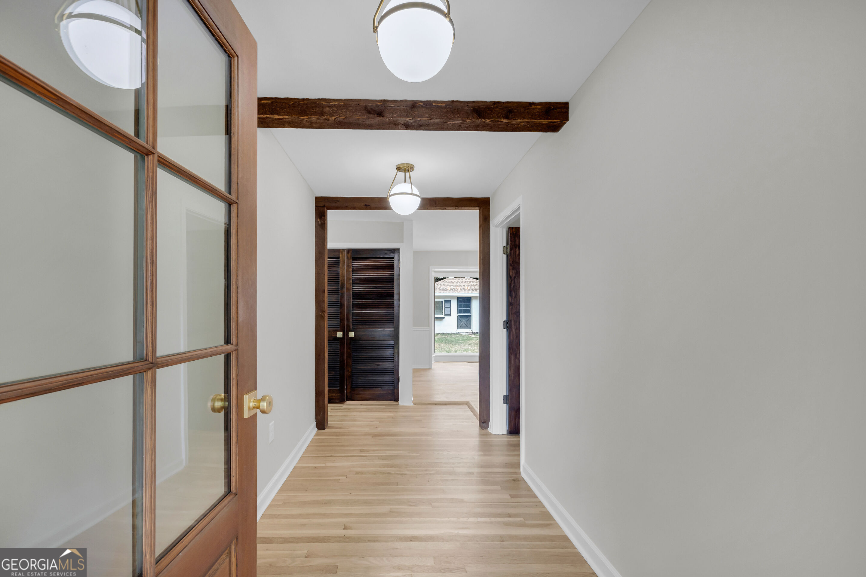 Empty hallway with light wood floors, exposed beams, and an open wood-framed glass door.
