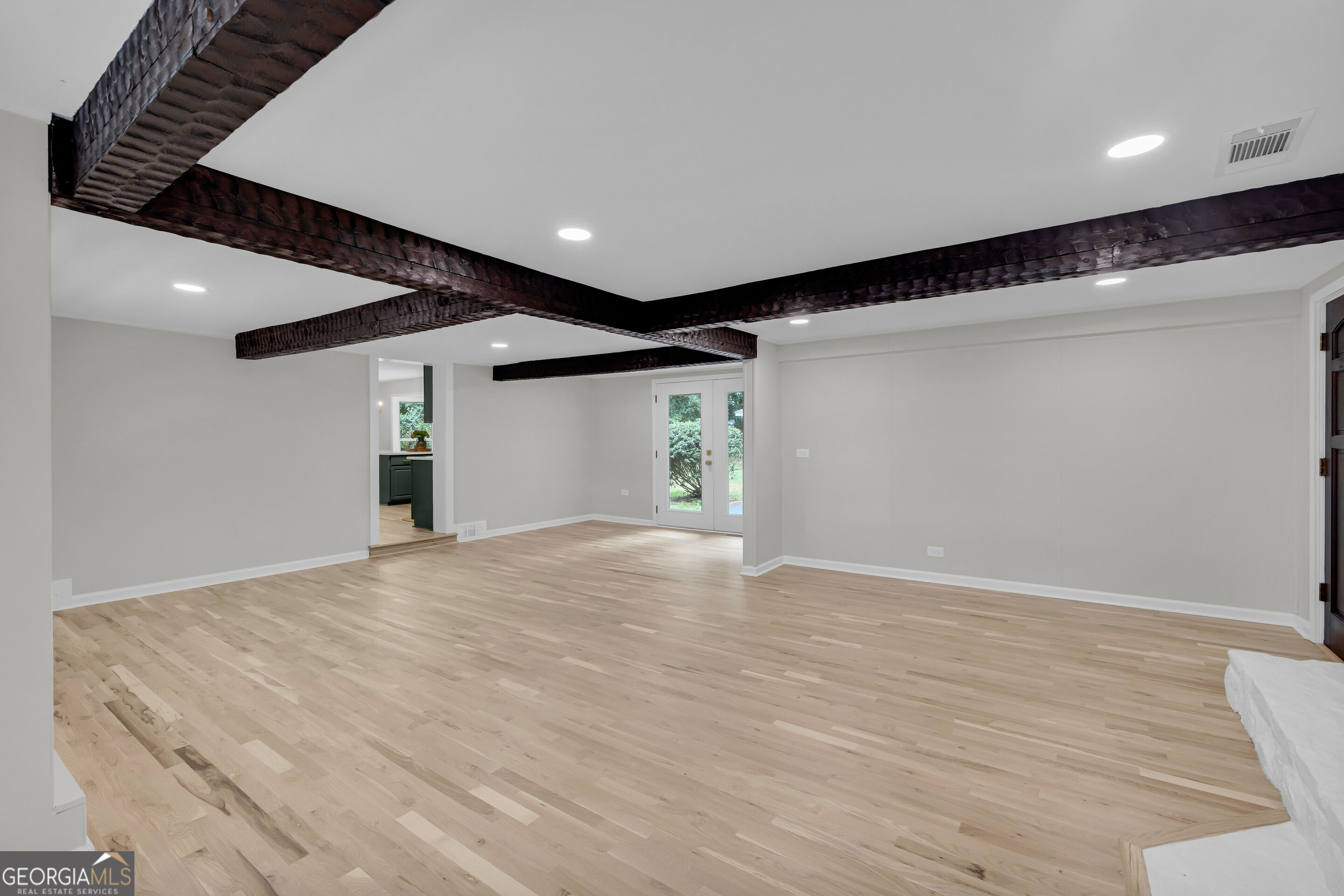 Vacant living room with pale wood floors, dark exposed ceiling beams, and French doors to greenery.