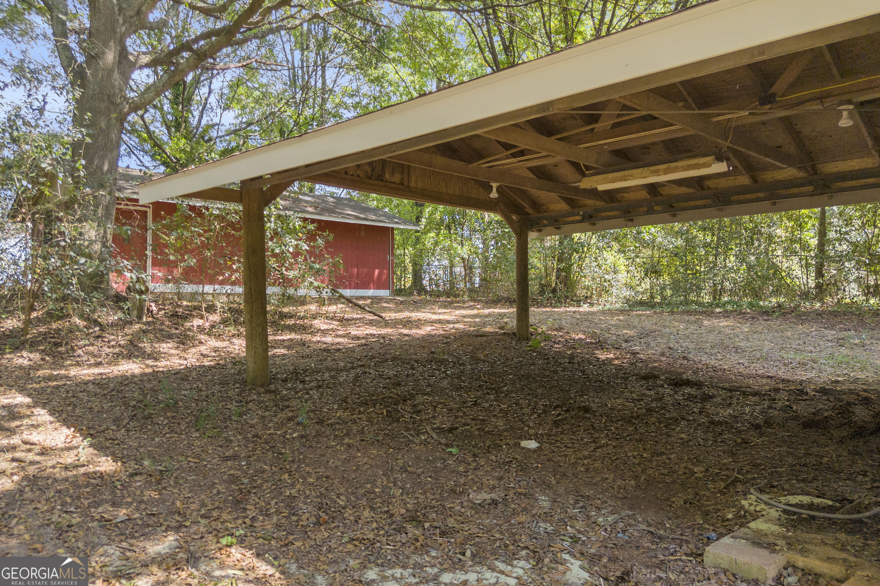Covered wooden carport among trees, with a red outbuilding behind it.