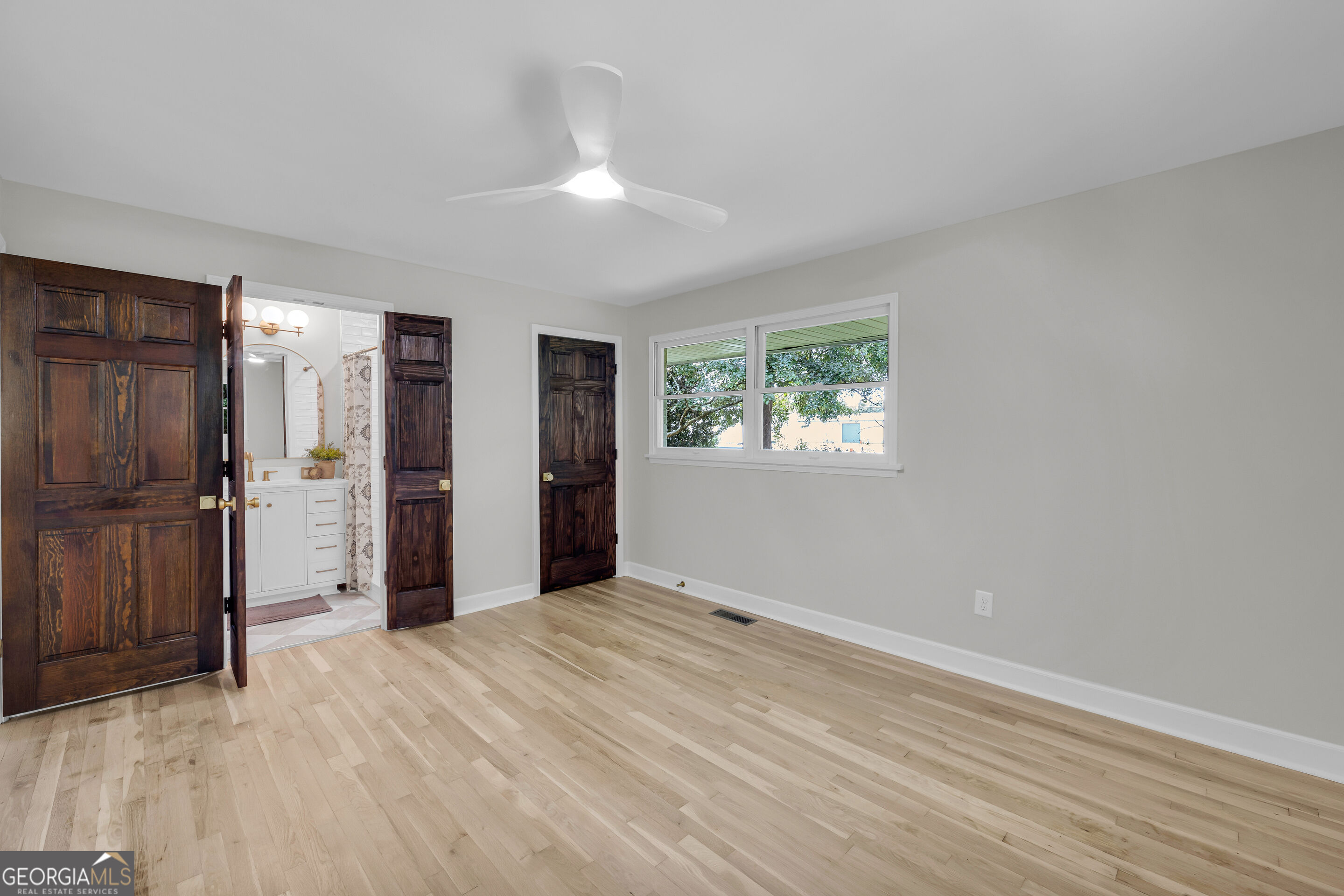 Empty bedroom with light hardwood floors, dark wood doors, and an adjoining bathroom.