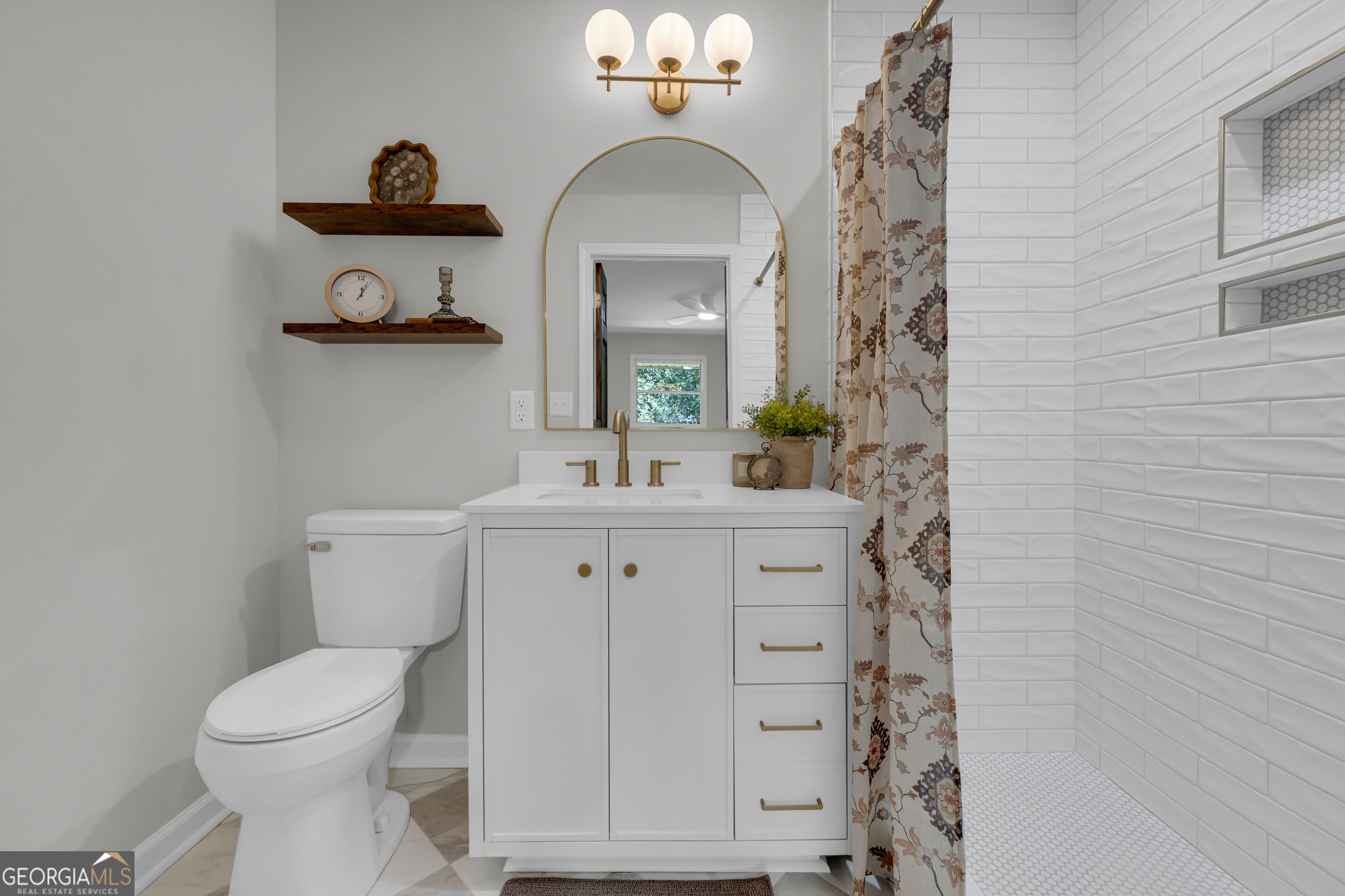 Bathroom with white vanity, brass fixtures, subway-tile shower, and patterned curtain.