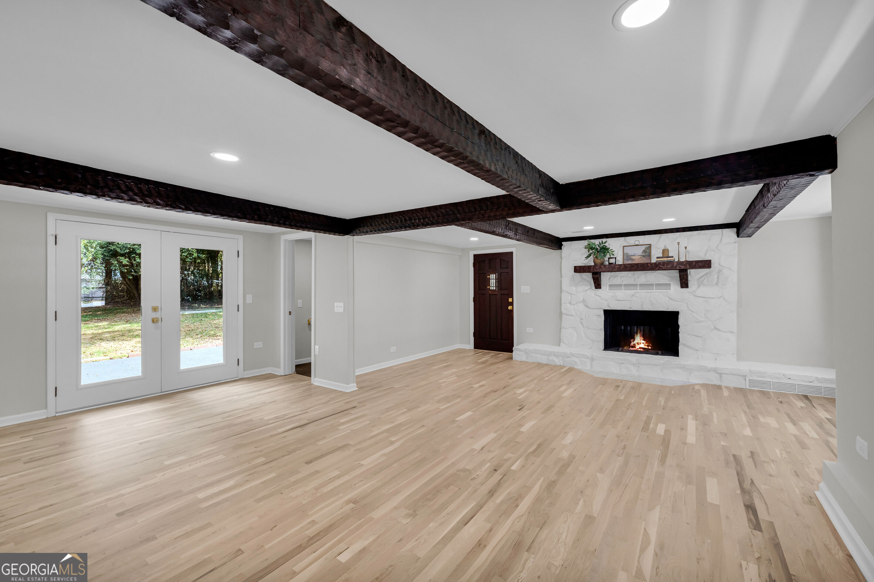 Vacant living room with exposed dark beams, light hardwood floors, and a white stone fireplace.