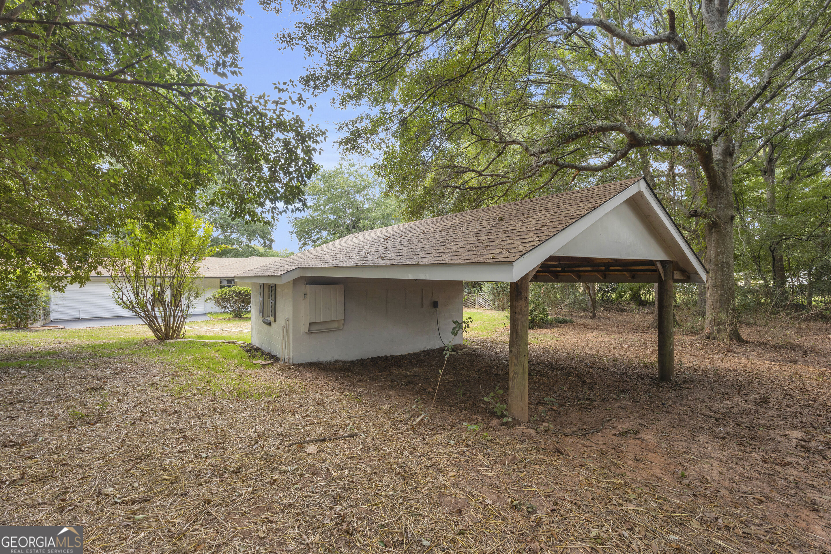 Covered carport beside a light-colored building, surrounded by mature trees and a leaf-covered yard.