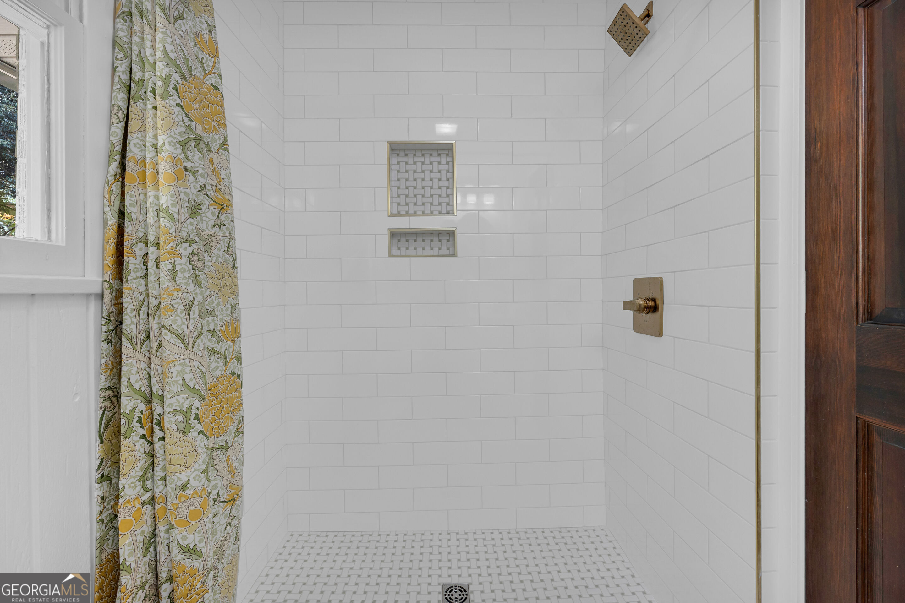 Walk-in shower with white subway tile, brass fixtures, recessed niches, and a floral curtain.