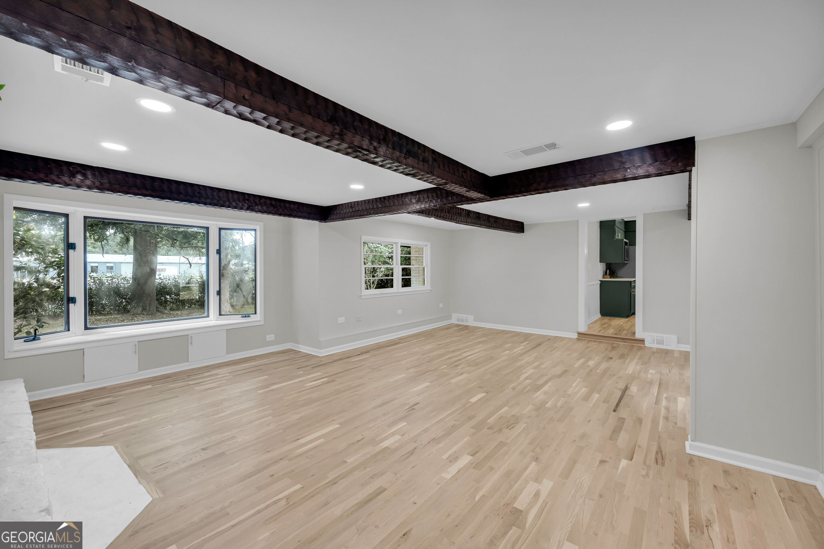Empty living room with exposed dark ceiling beams, light wood floors, and large windows.