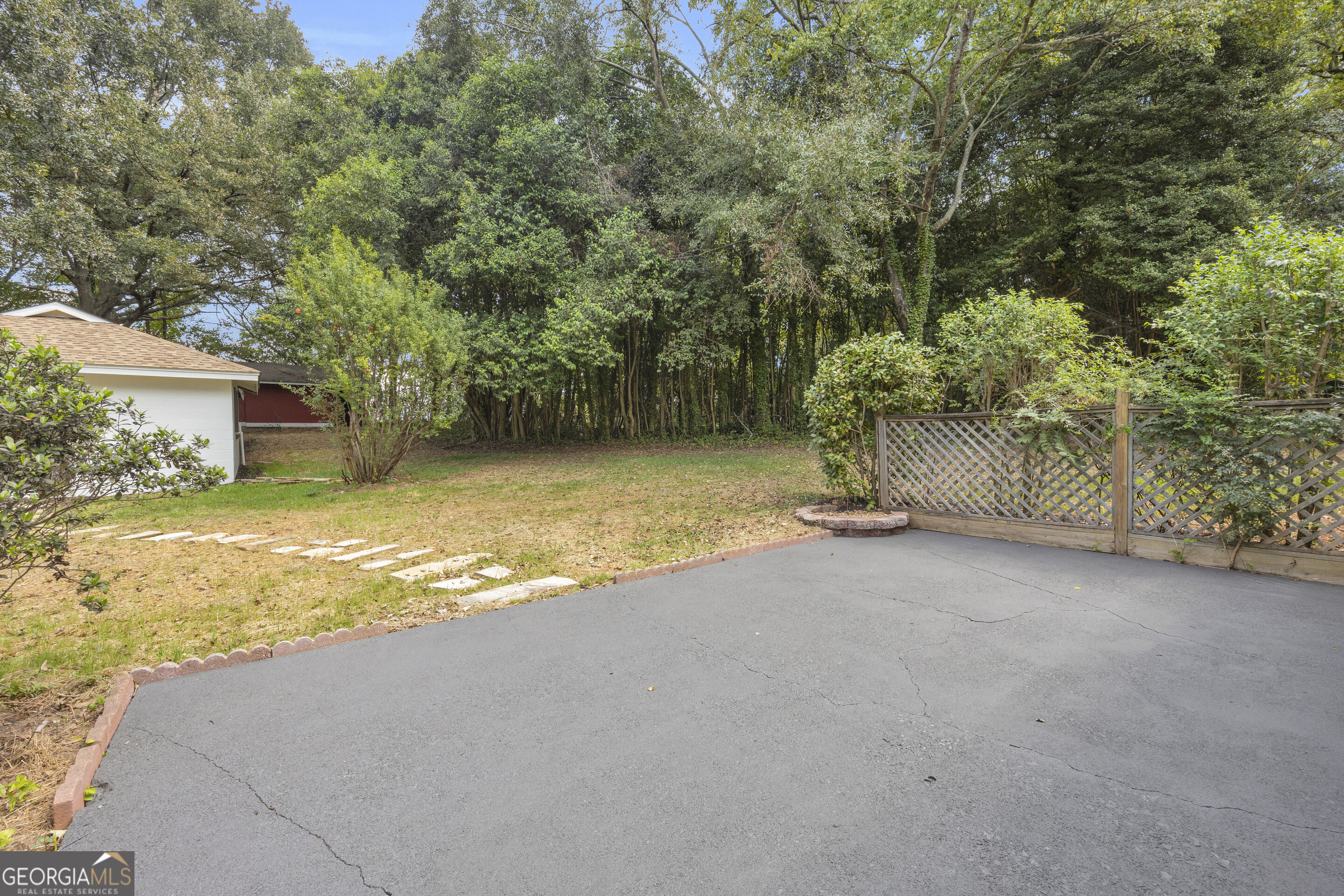Paved backyard beside a lawn, lattice fence, stepping stones, and mature trees.