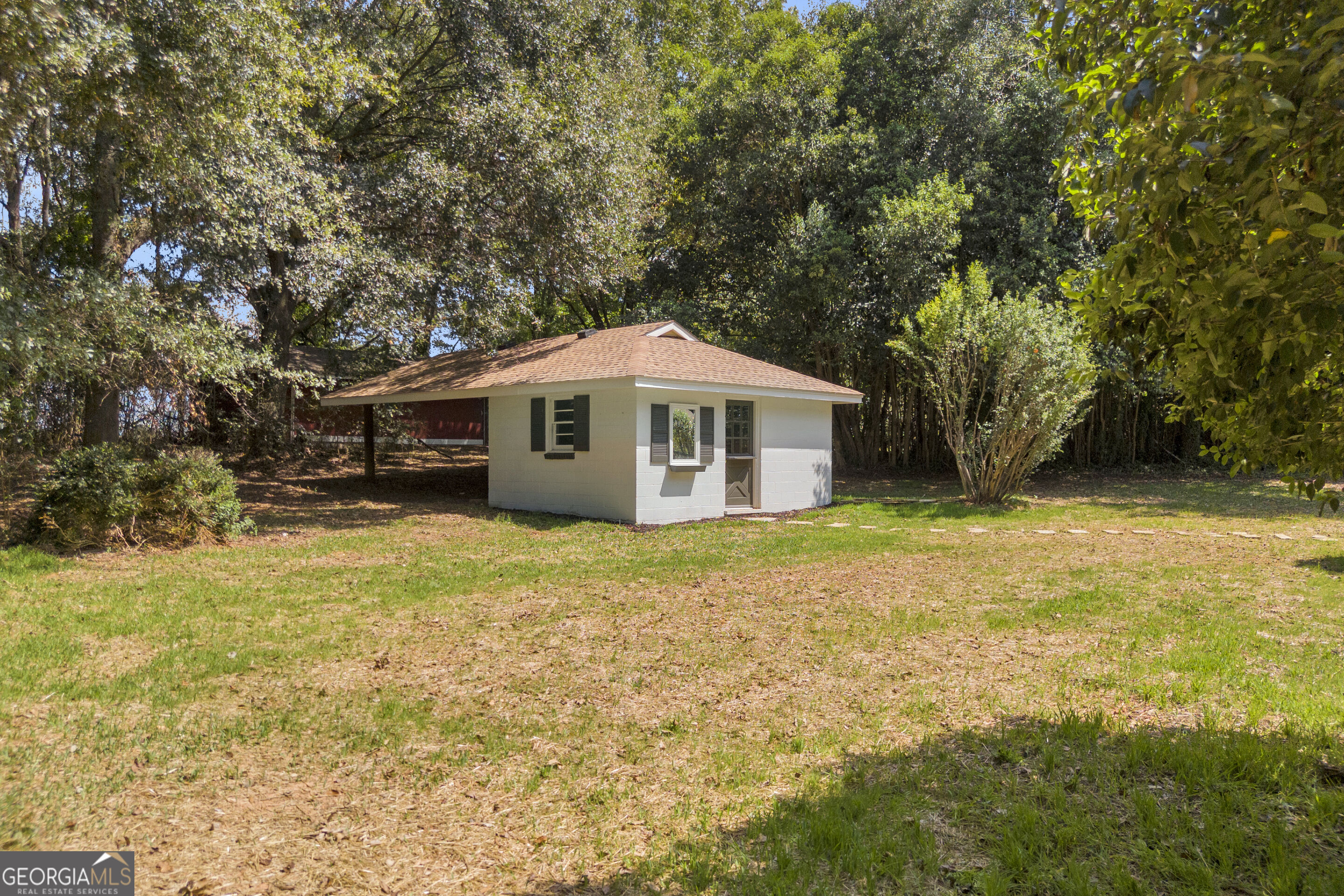 Small light-colored outbuilding with a reddish-brown roof in a grassy, wooded yard.
