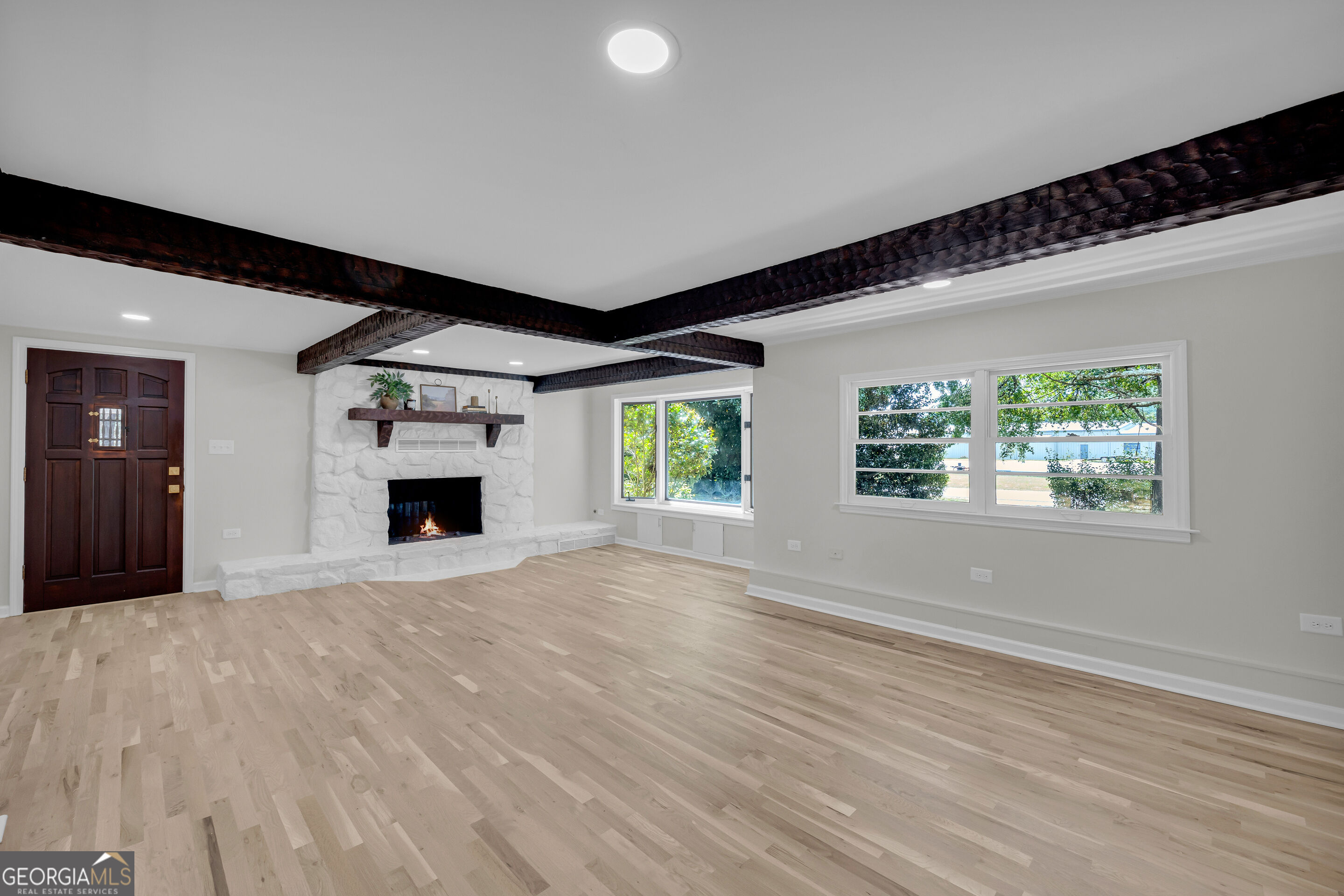Empty living room with pale wood floors, dark ceiling beams, and a white stone fireplace.