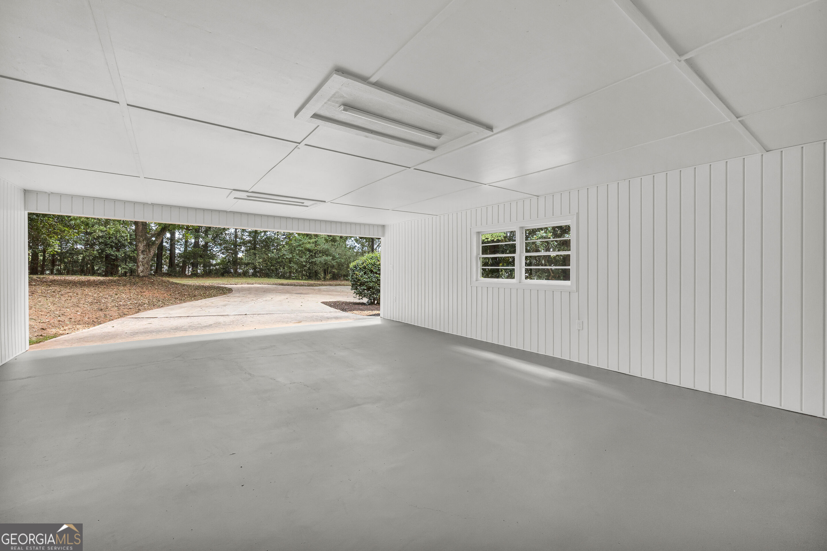 Empty white-paneled garage with a concrete floor, wide open side, and view of a wooded yard.