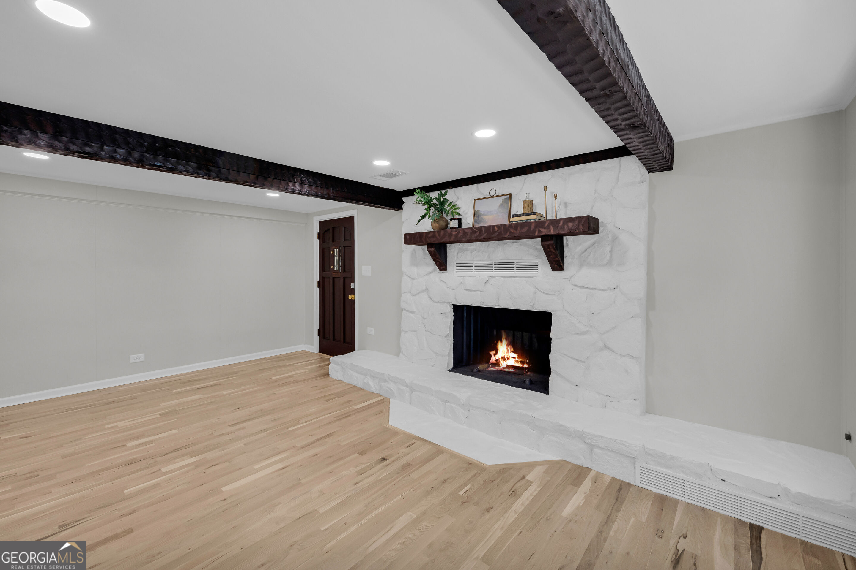 Empty living room with a white stone fireplace, burning fire, dark ceiling beams, and pale wood floors.