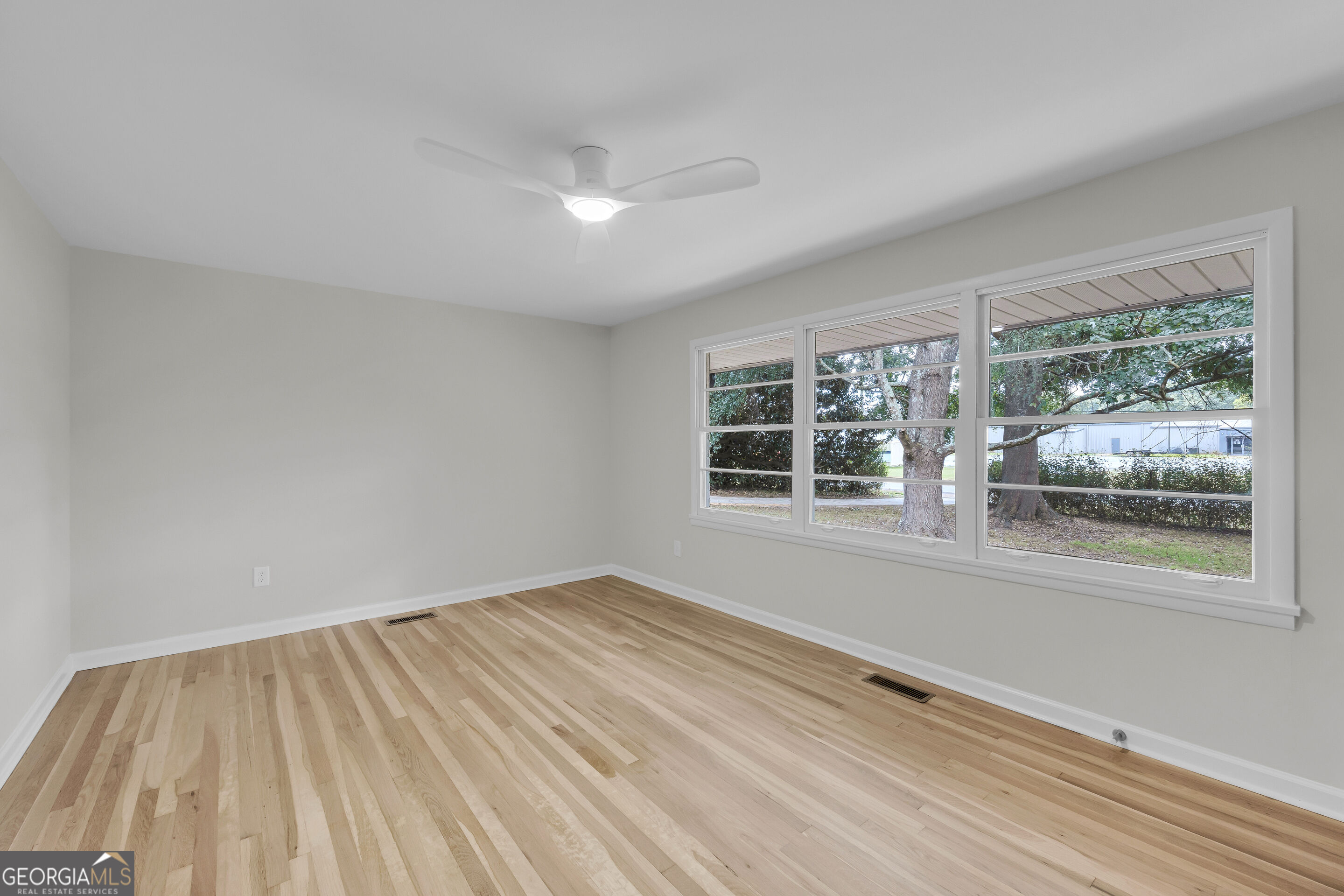 Empty room with light hardwood floors, ceiling fan, and wide windows overlooking trees.