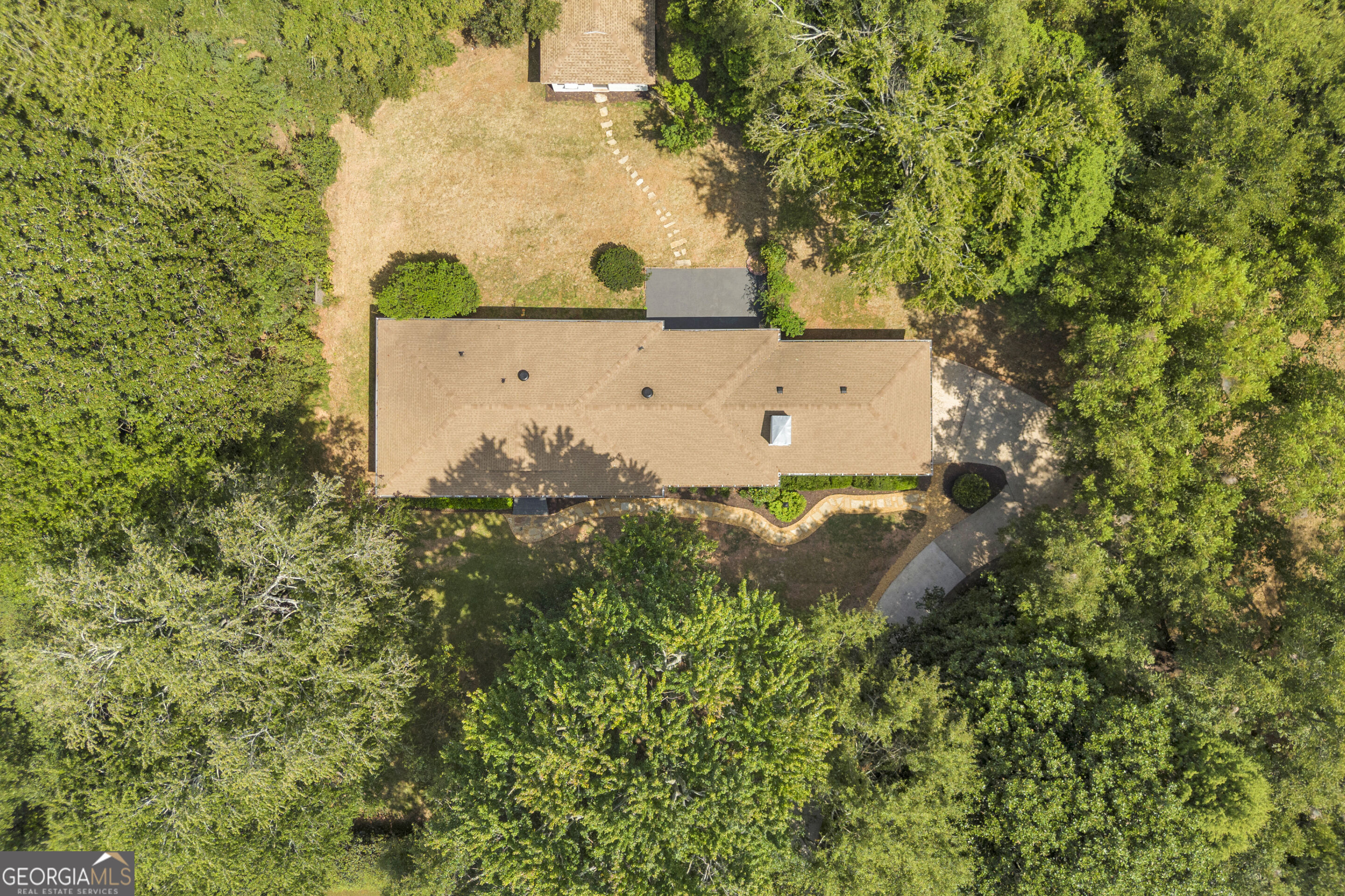 Aerial view of a tan-roofed home surrounded by mature trees, with a curved driveway and landscaped yard.