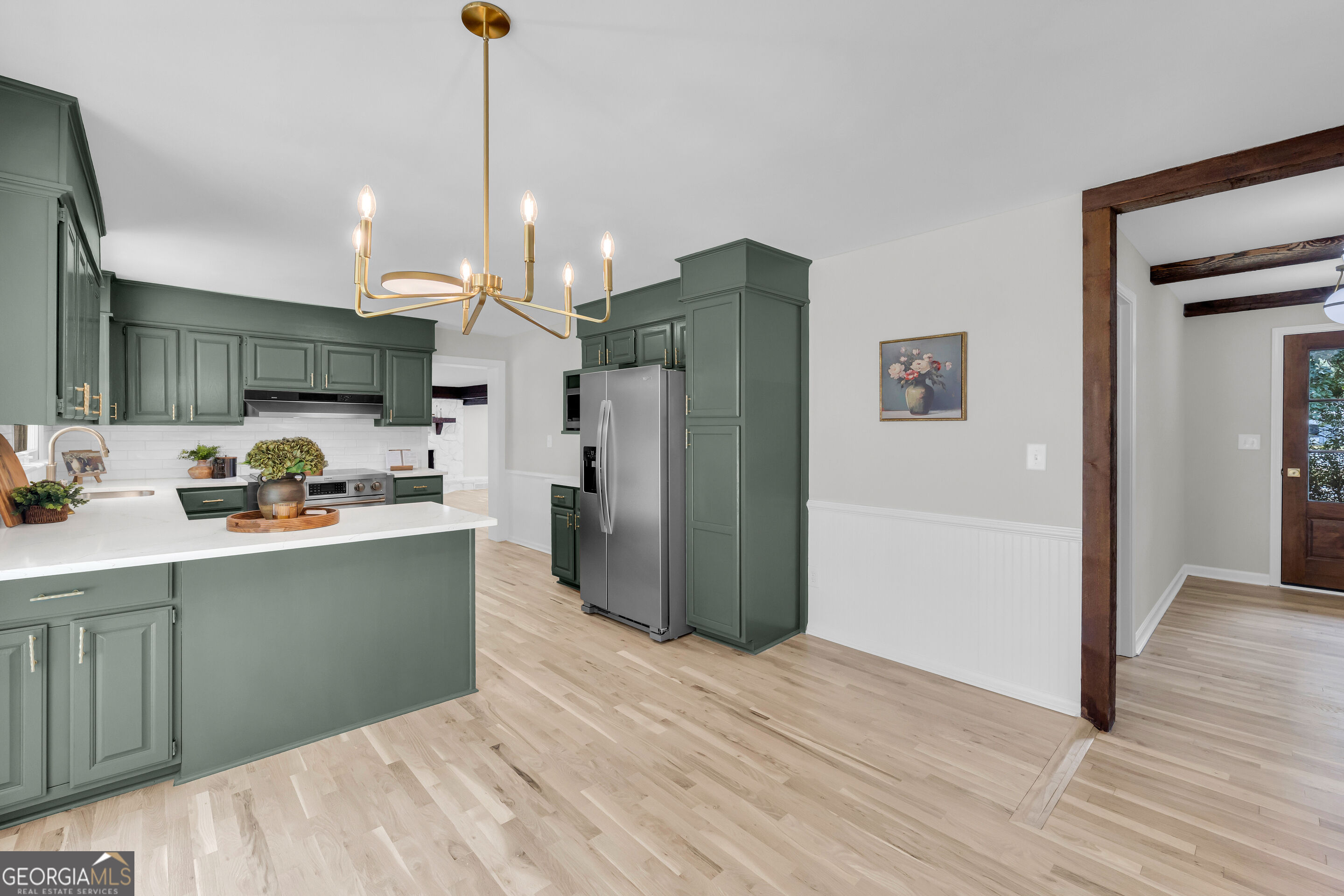 Kitchen with deep green cabinets, a white island countertop, stainless steel refrigerator, and brass chandelier.