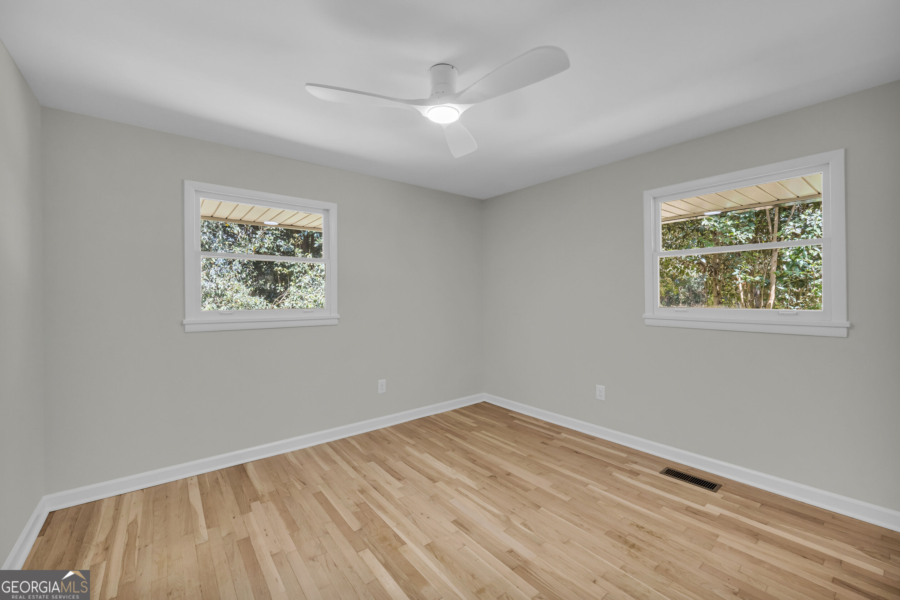 Empty room with light hardwood floors, two windows overlooking trees, and a ceiling fan.