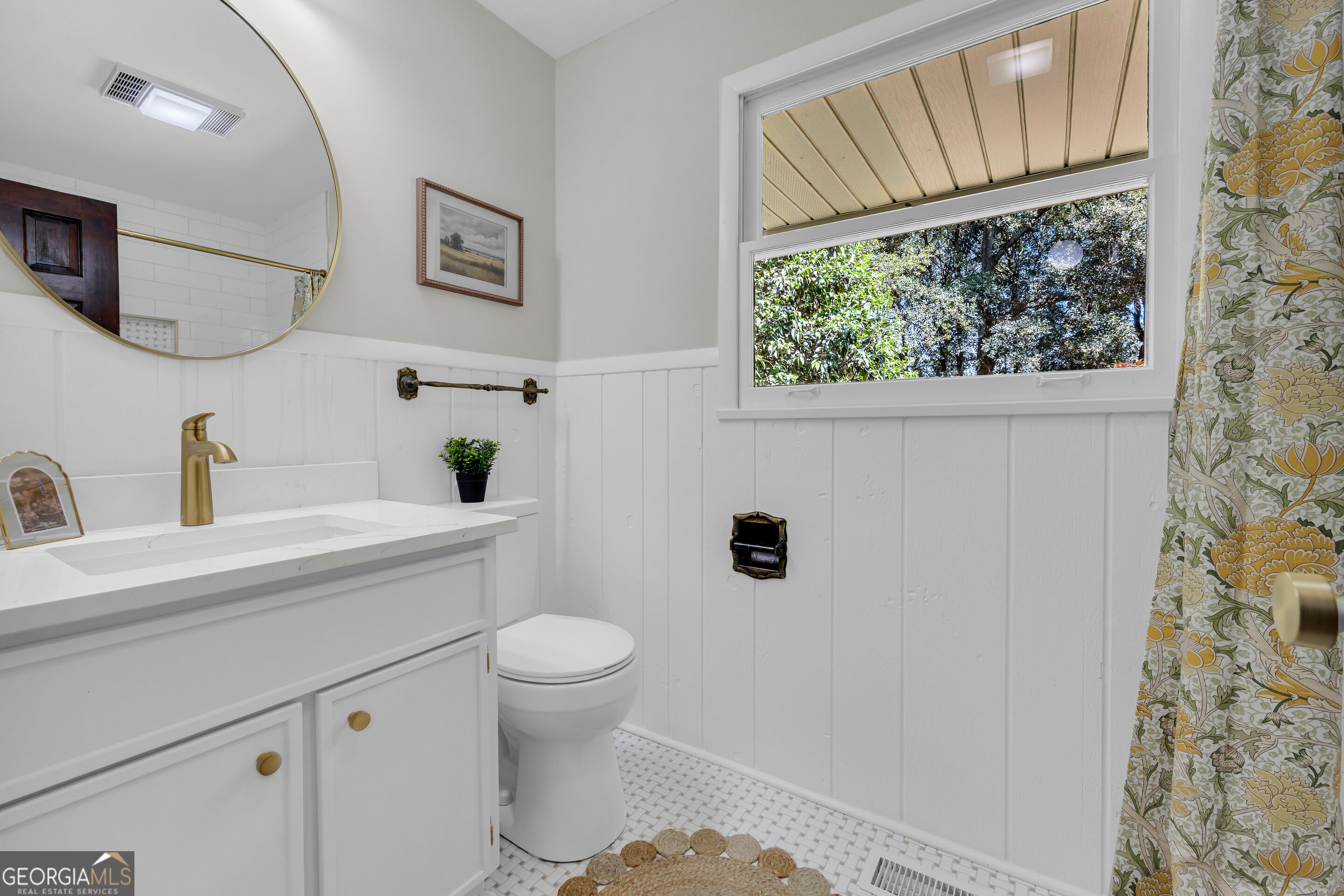White bathroom with a gold faucet, botanical shower curtain, and window overlooking trees.