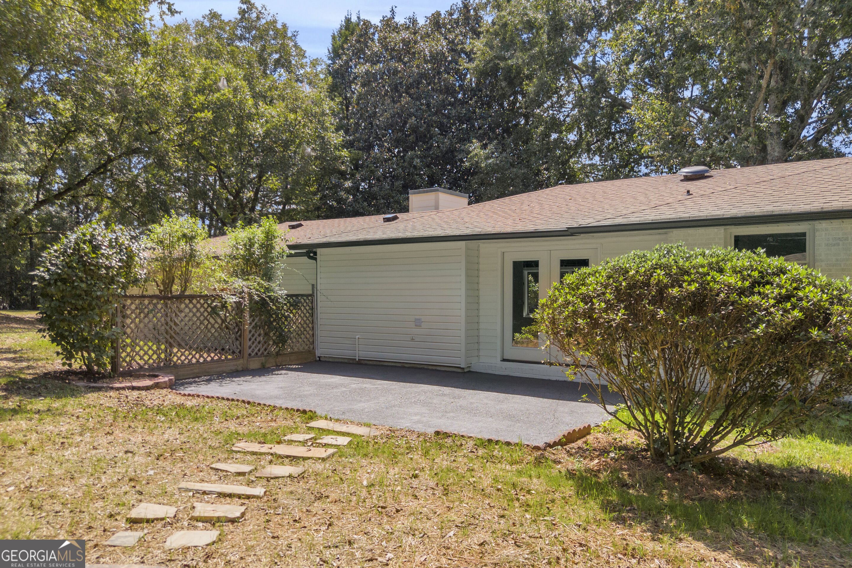 Rear of a single-story house with a paved patio, double doors, and mature shrubs.