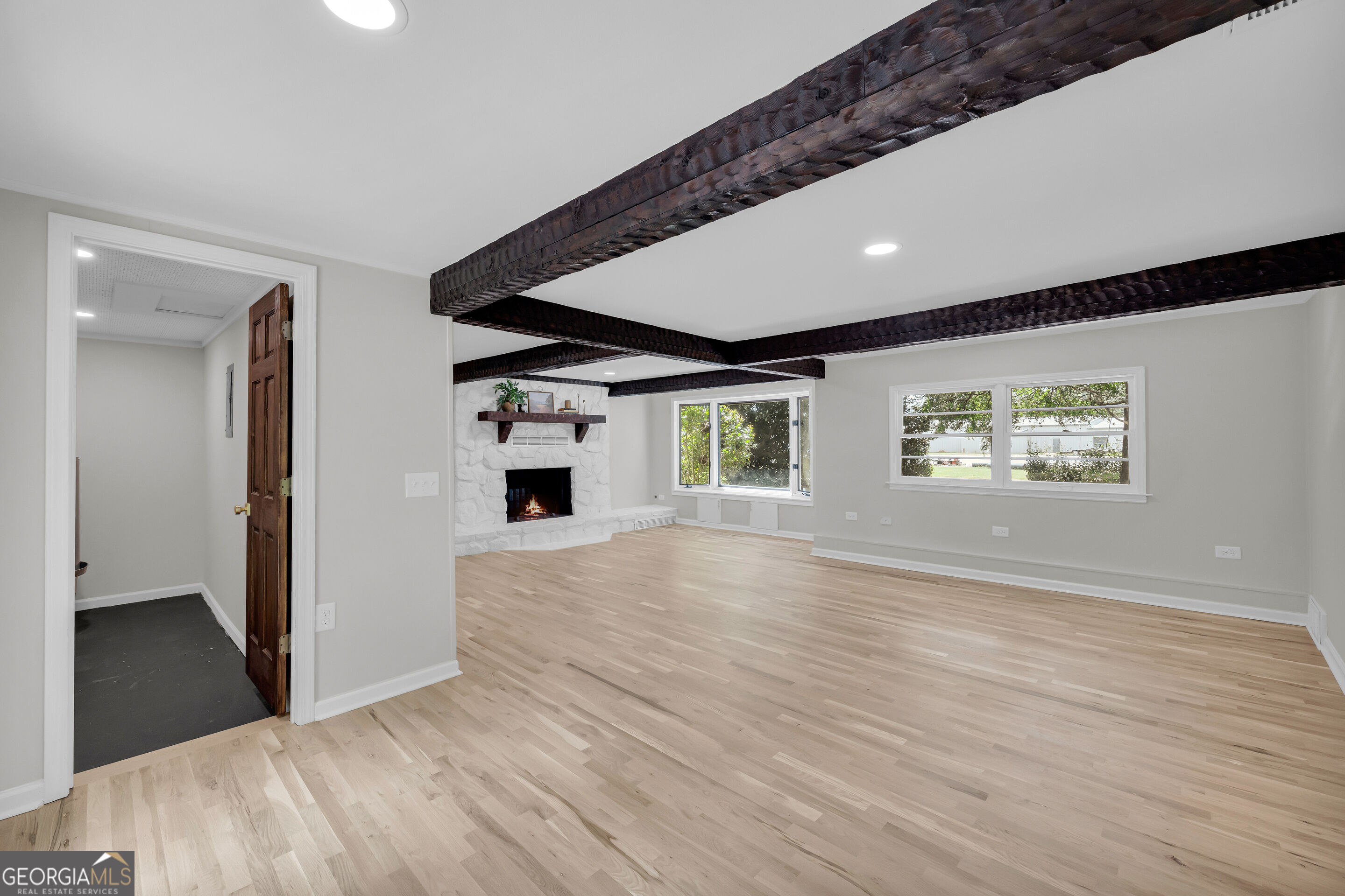 Empty living room with dark exposed ceiling beams, pale wood floors, and a white stone fireplace.