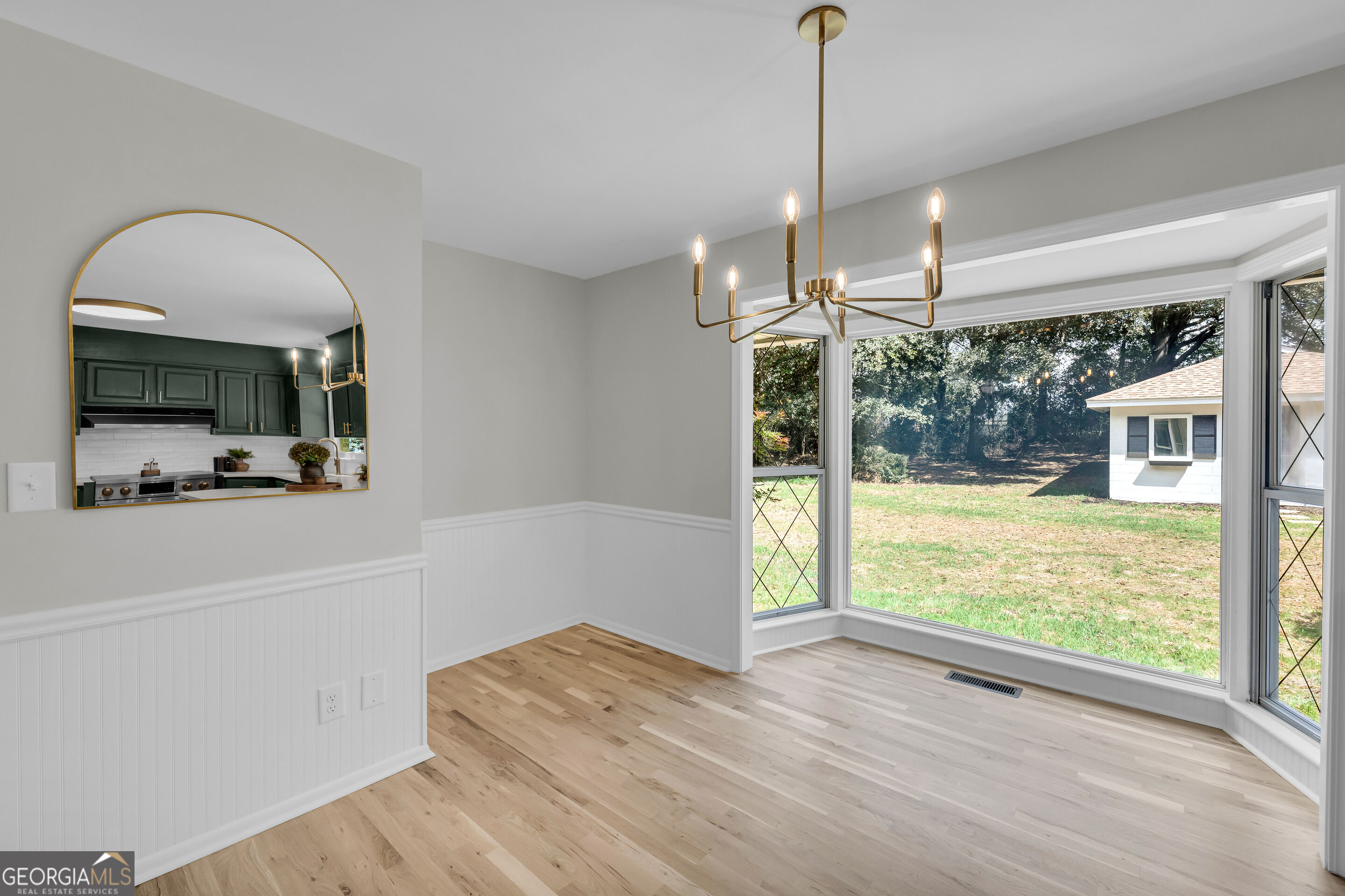 Dining area with a brass chandelier, large yard-facing windows, and an arched mirror reflecting the kitchen.